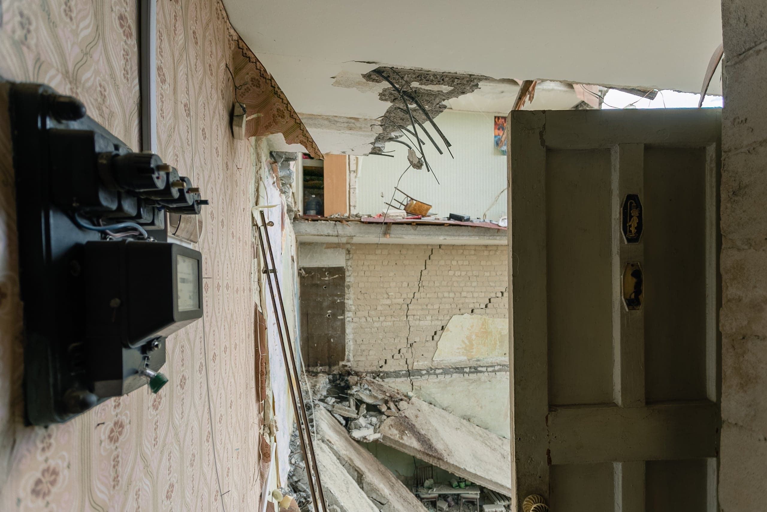 Взгляд из разрушенного помещения с электросчетчиком на стене на обломки и поврежденный фасад здания напротив.