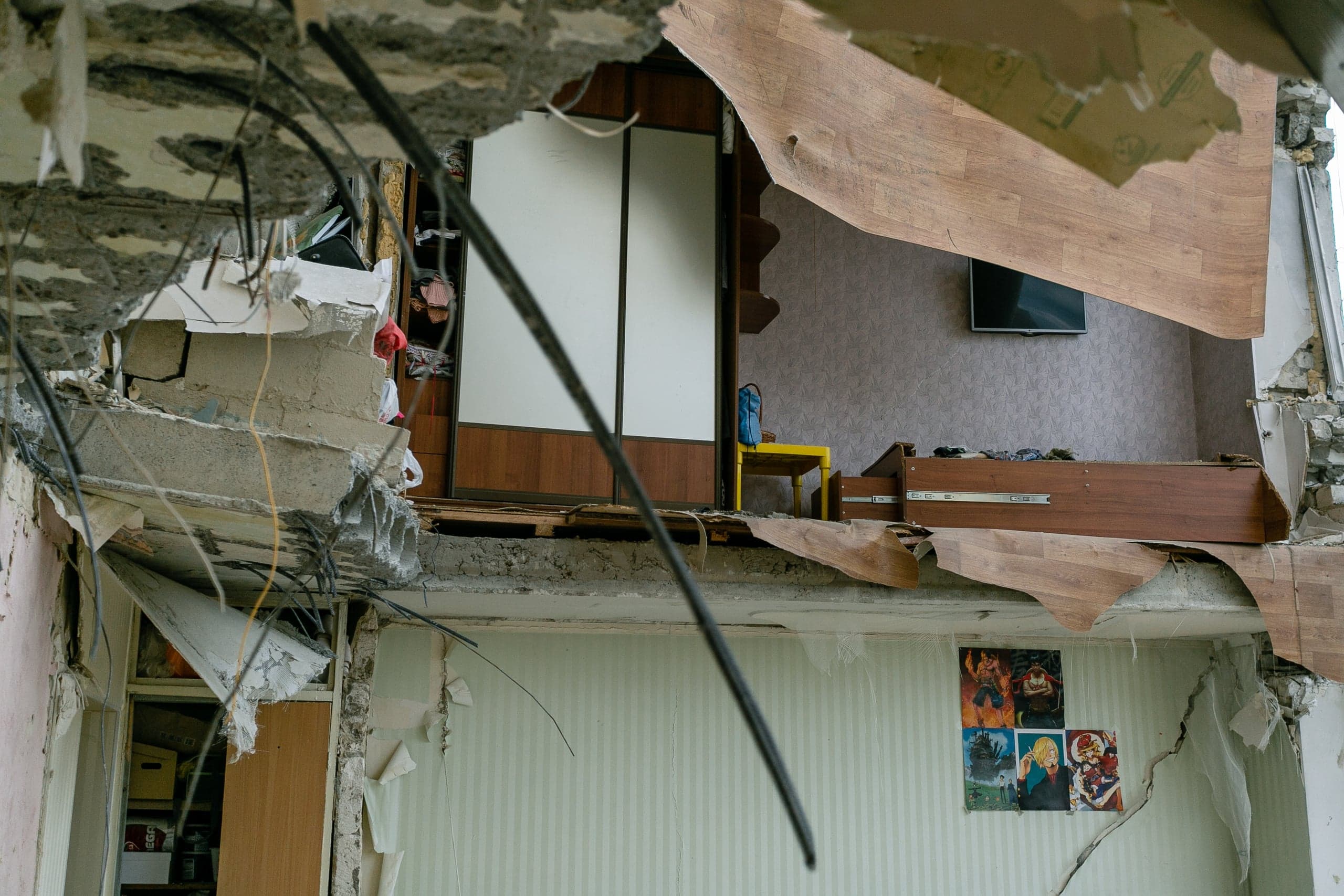 Разрушенный интерьер жилого дома: вид сквозь пролом в потолке с обнаженной арматурой на уцелевшую мебель и плакаты на стене.