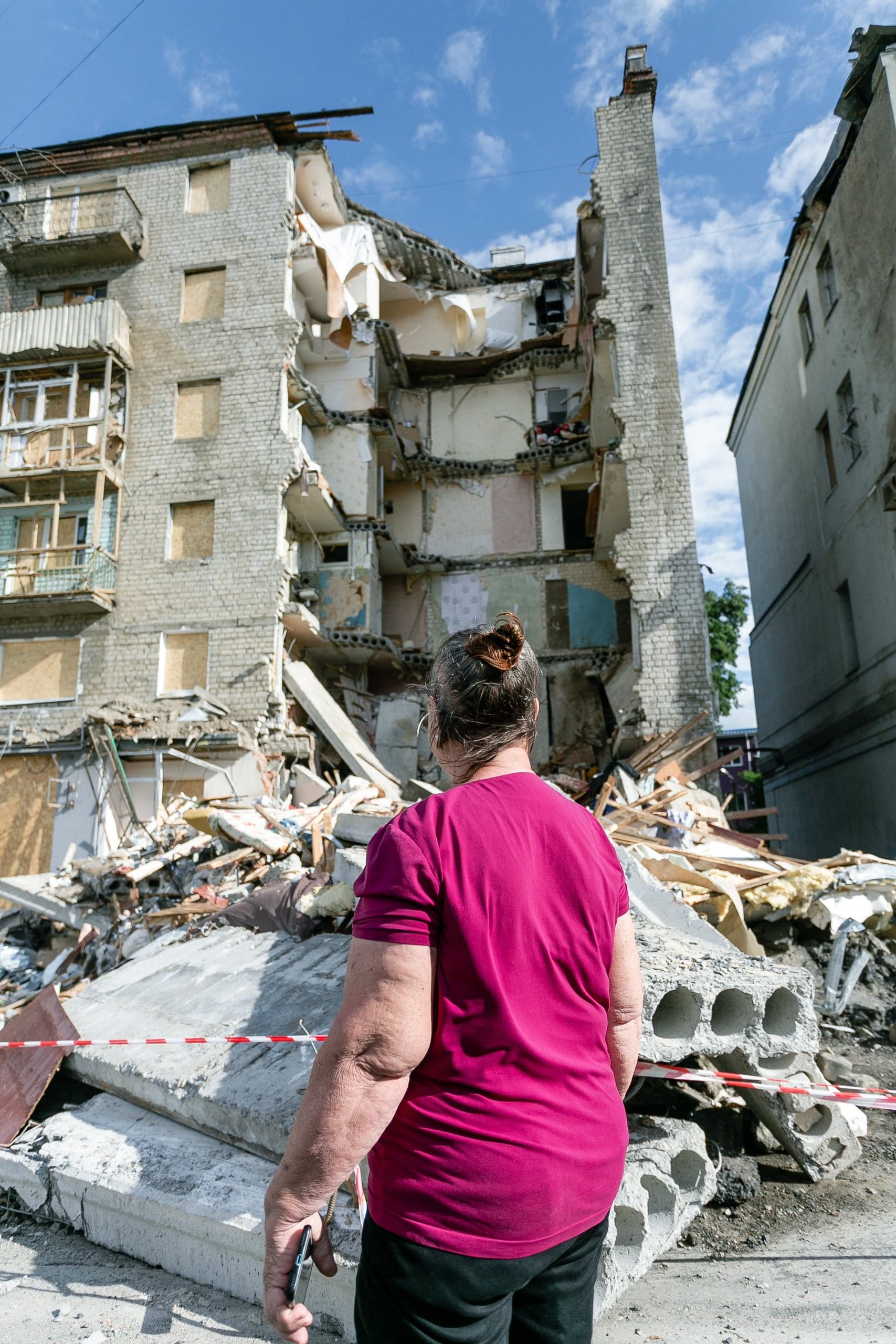 Женщина стоит спиной, глядя на разрушенный многоэтажный дом и груды обломков под голубым небом.