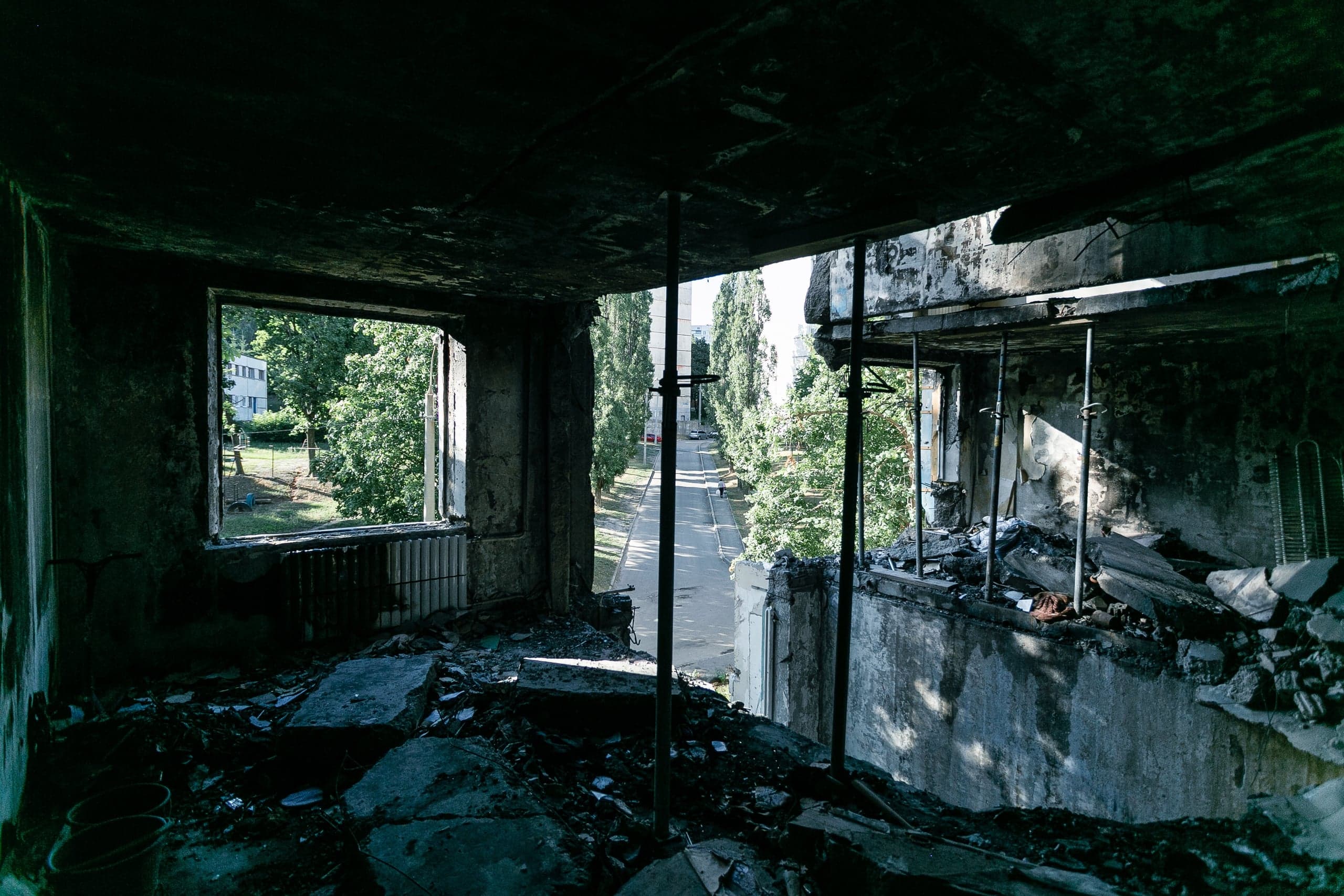 Обгоревший интерьер разрушенной квартиры с обломками. Сквозь проемы видна зеленая улица, обсаженная высокими деревьями.