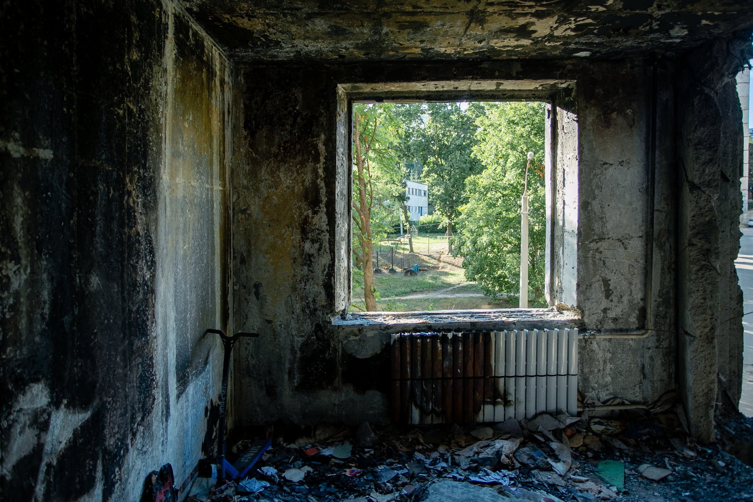 Интерьер сильно обгоревшей комнаты с черными стенами и мусором на полу. Через пустой оконный проем виден зеленый двор и деревья.