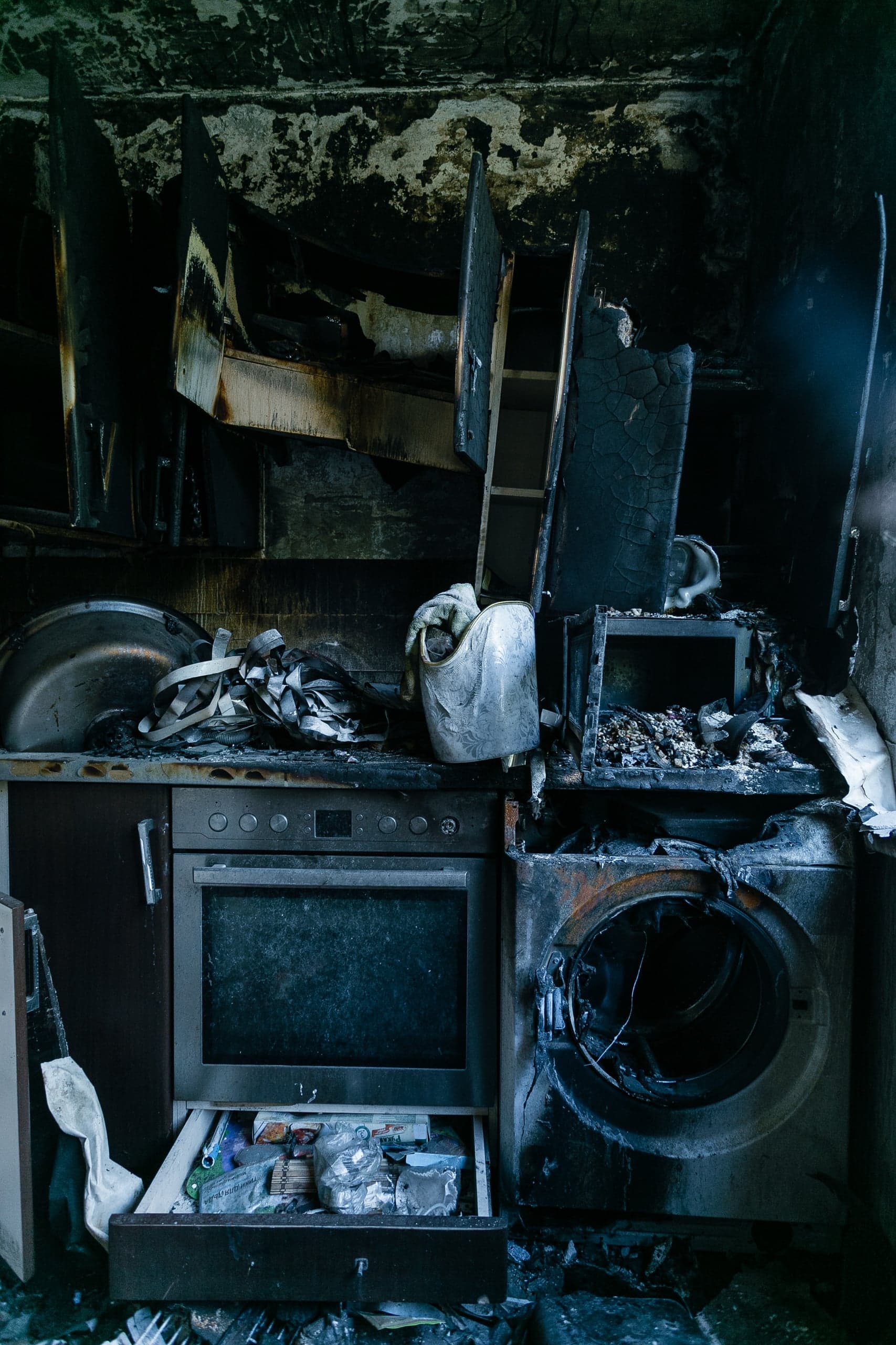 Сгоревшая, закопченная кухня: обугленные шкафы, духовка, стиральная машина и открытый ящик с предметами.
