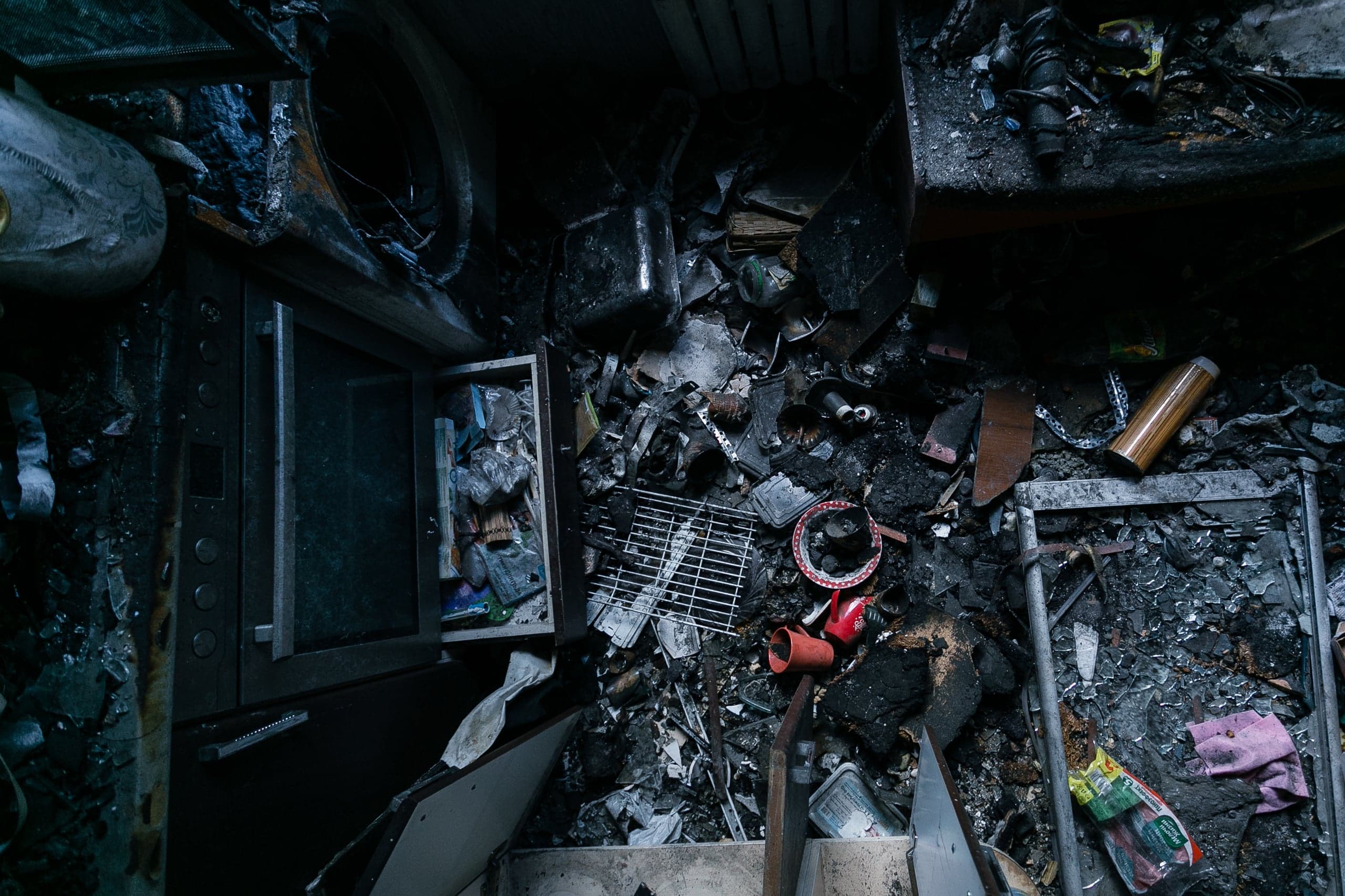 Сгоревший интерьер комнаты, заваленной обломками, пеплом и оплавленной бытовой техникой.