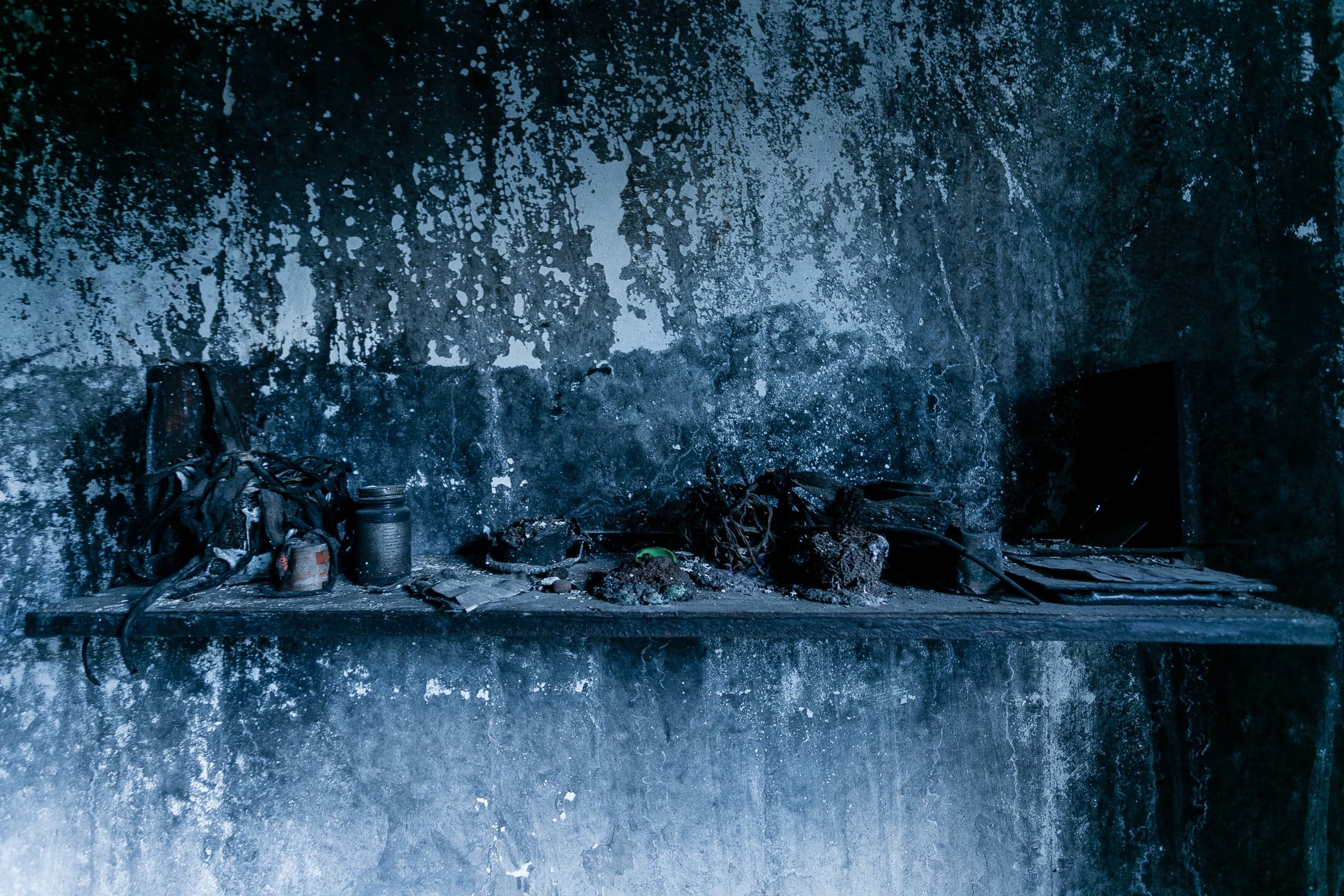 Обугленная стена с облупившейся краской и полка с мусором и банками в темно-синих тонах.
