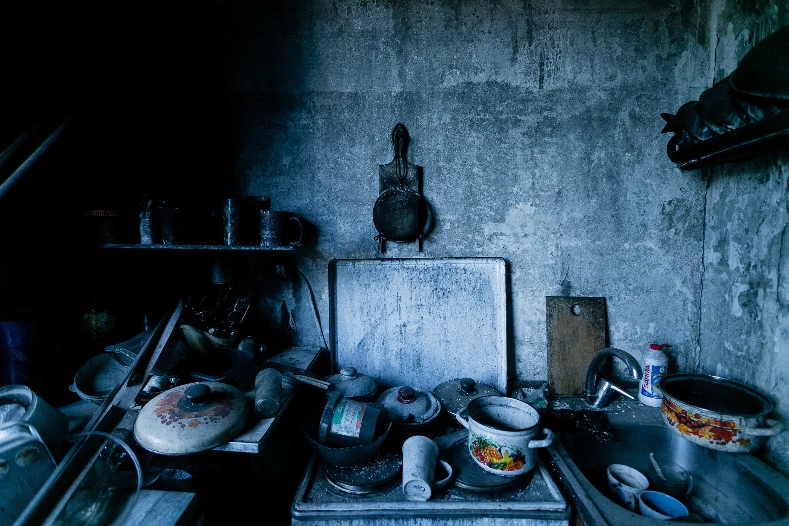 Заброшенная, захламленная кухня. Столешница и плита, заваленные грязной посудой и утварью. Обветшалые сине-серые бетонные стены.