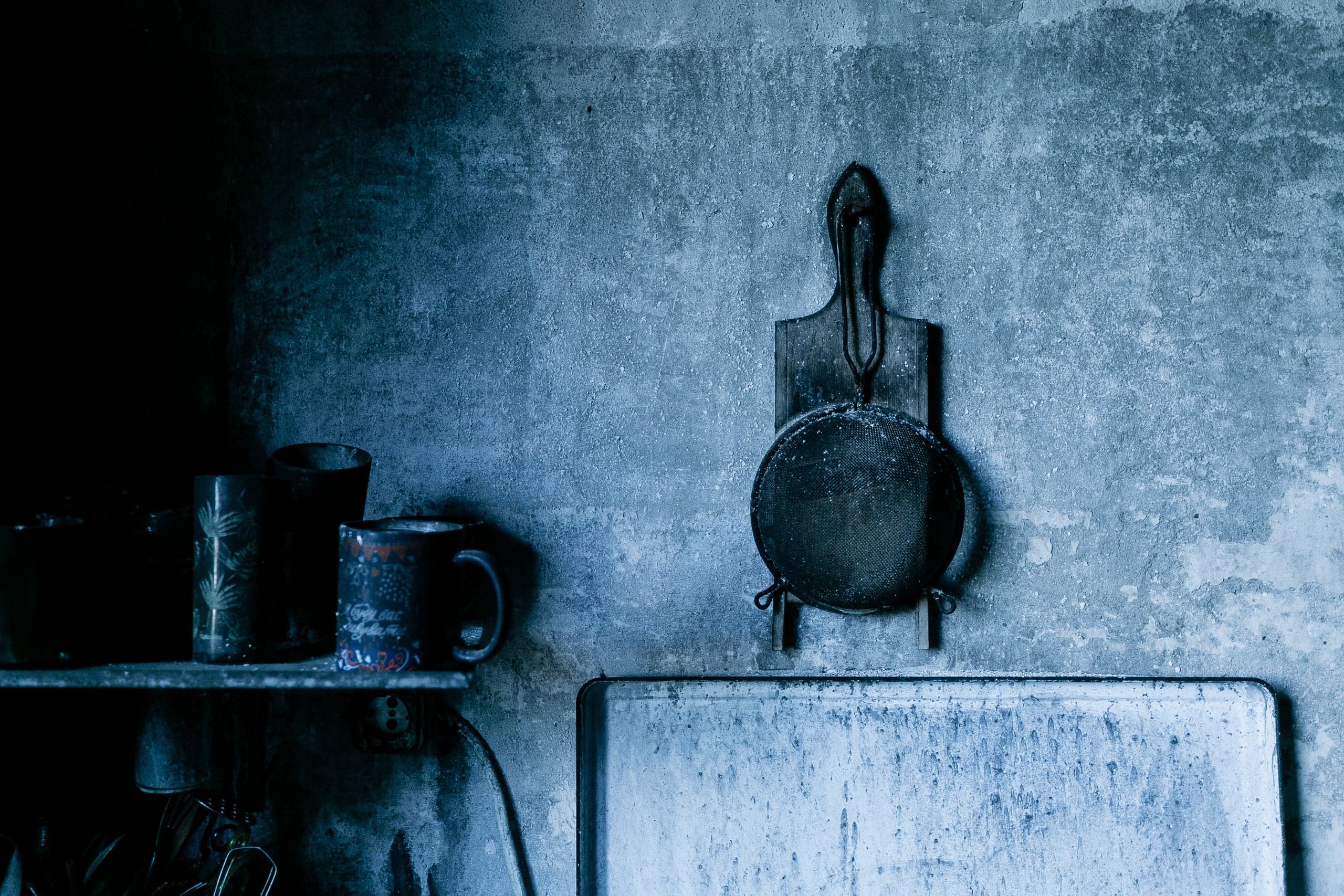 Мрачный натюрморт в синих тонах: сито и разделочная доска на текстурной стене, рядом кружки на полке.