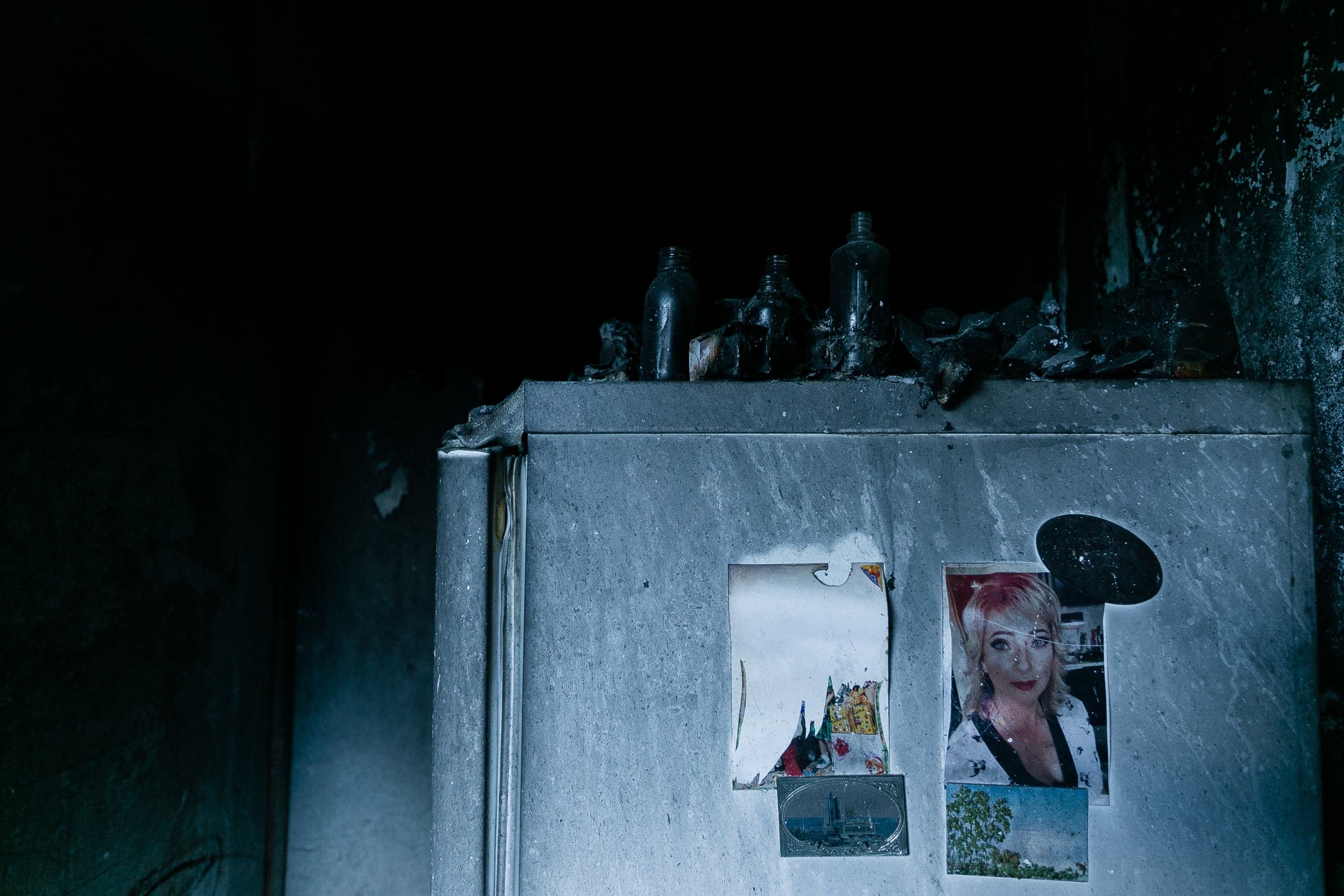 Обгоревший шкаф в темном, закопченном помещении. На шкафу — оплавленные бутылки и приклеенные фотографии, включая портрет улыбающейся женщины.