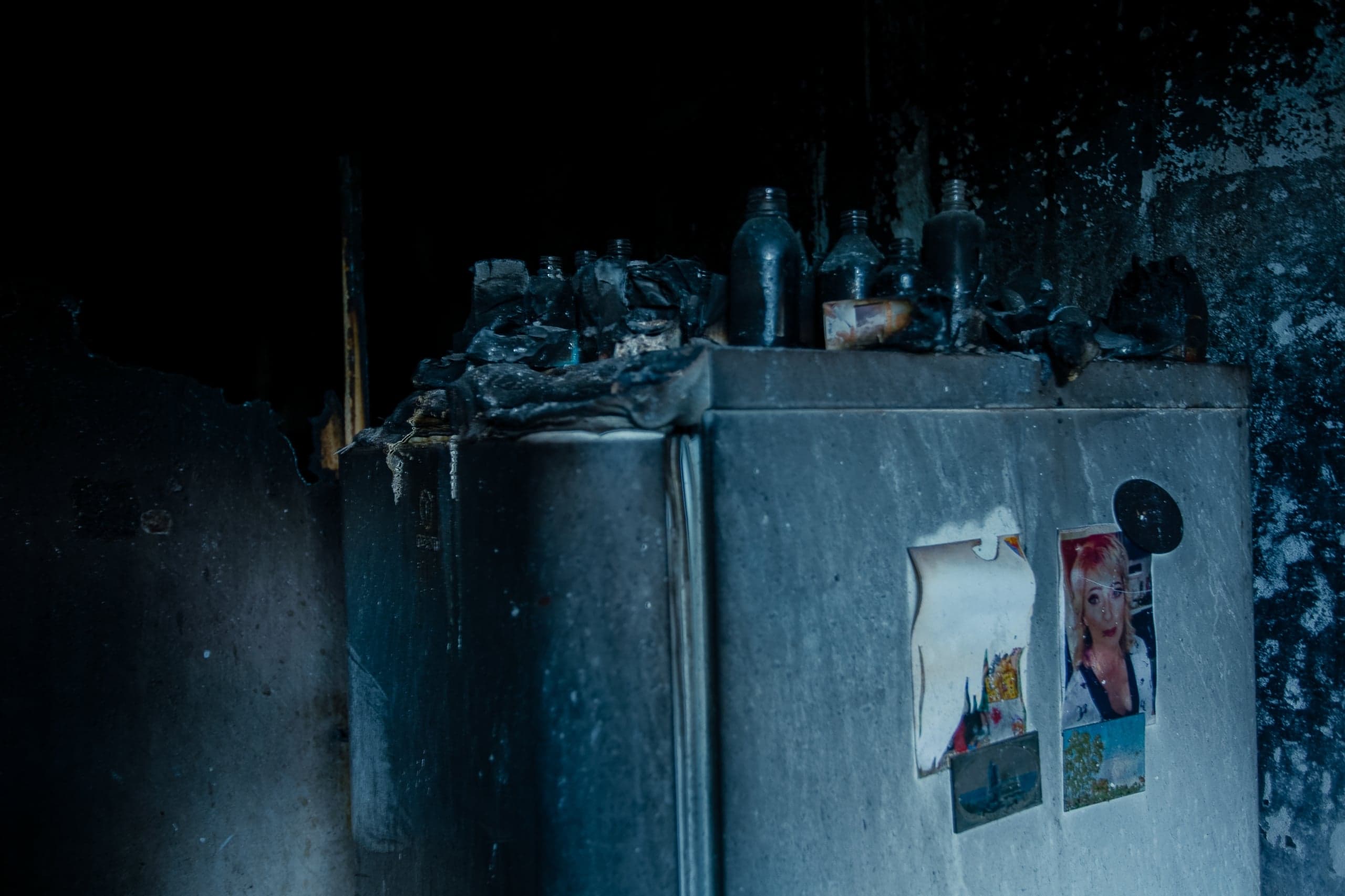 Закопченный холодильник после пожара. Сверху — расплавленные остатки и бутылки. На дверце — обгоревшие фотографии.