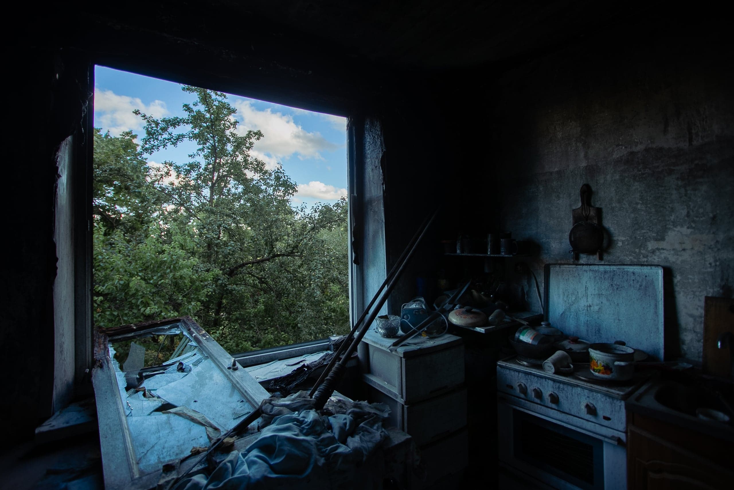 Темная, разрушенная комната (кухня) с разбитым окном. На переднем плане — кухонная плита и беспорядок, на заднем плане через окно виден яркий зеленый лес и небо.