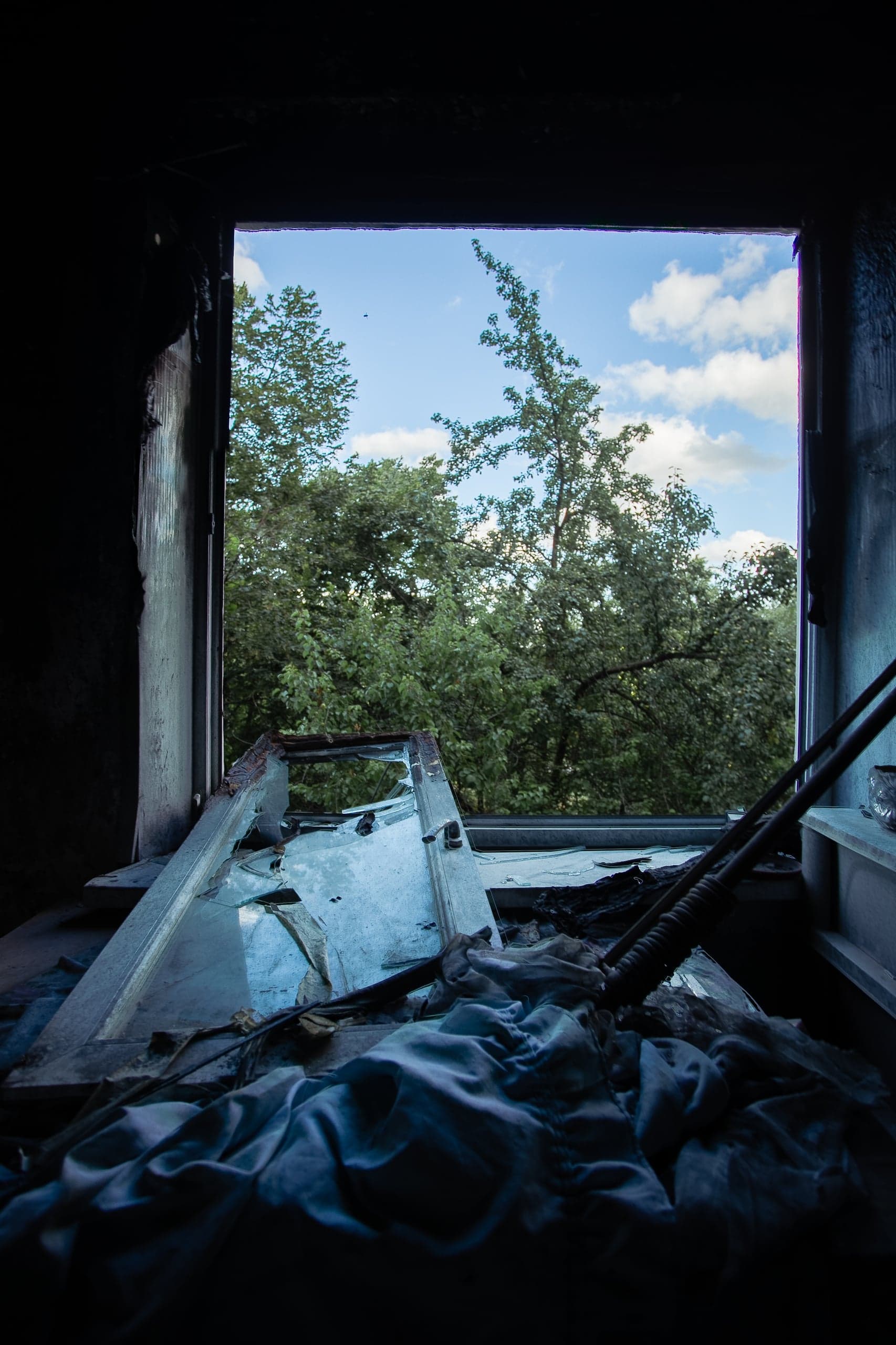 Оконный проем сгоревшего помещения с видом на зеленую листву и облачное небо; на подоконнике обломки и ткань.