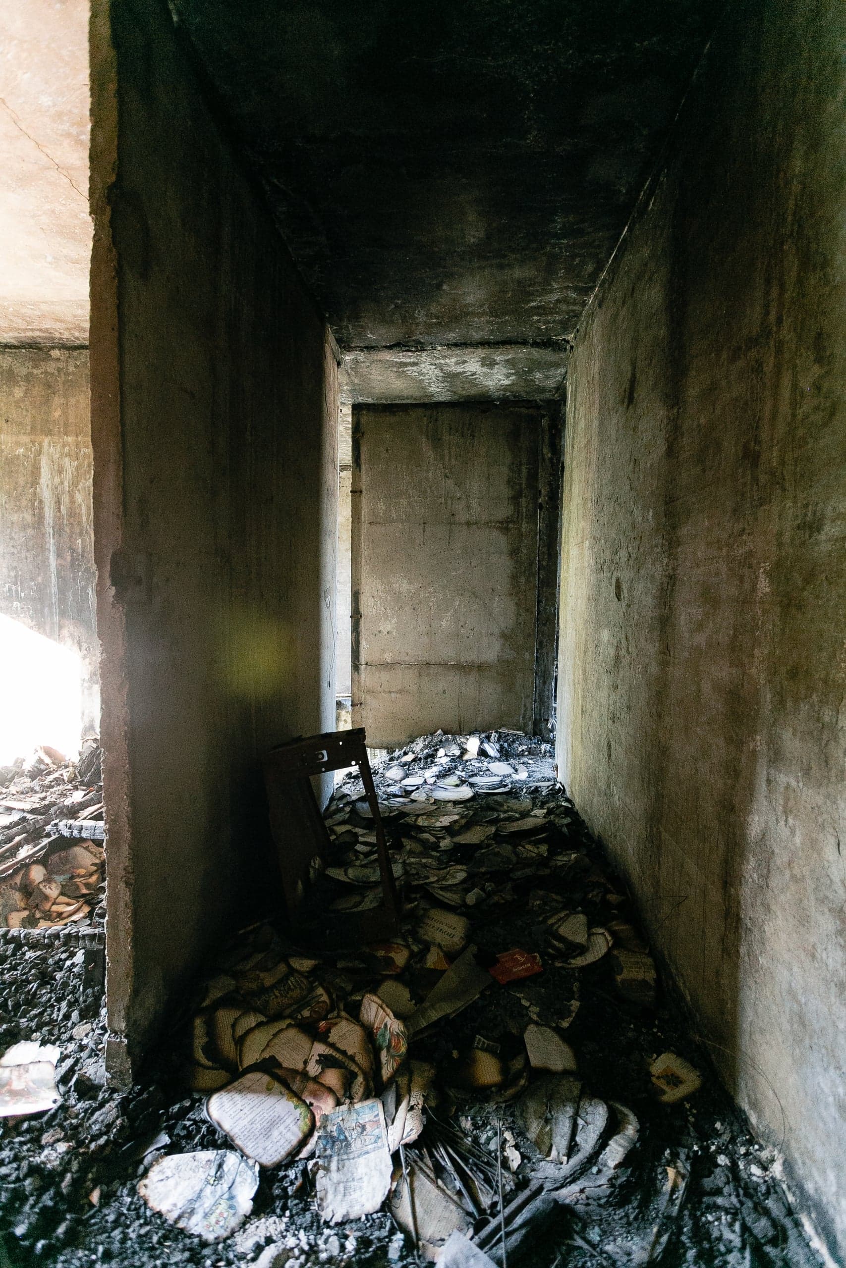 Узкий, темный коридор в разрушенном здании. Пол завален обломками и сгоревшими книгами. Контрастный свет.