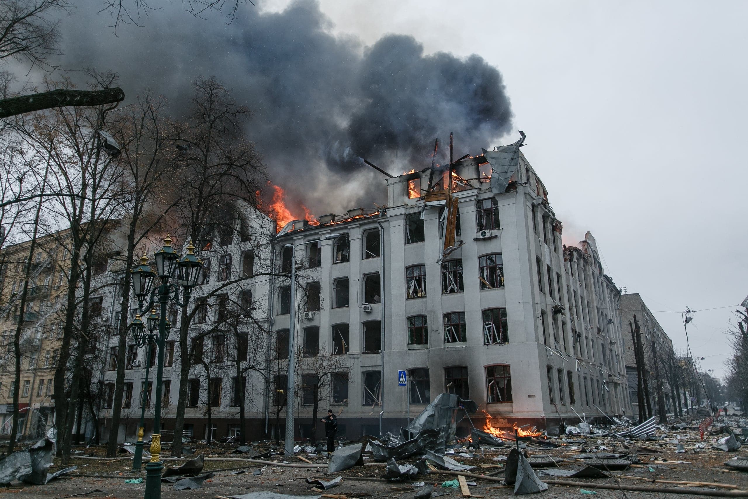Сильно поврежденное здание горит, густой черный дым поднимается с крыши. Фасад и улица усеяны обломками.
