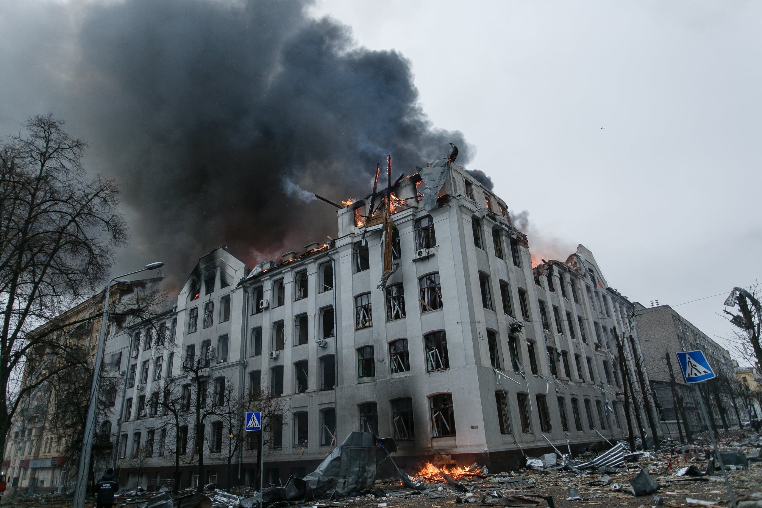 Горящее многоэтажное здание с выбитыми окнами. Над крышей поднимается столб густого черного дыма. Улица усеяна обломками.
