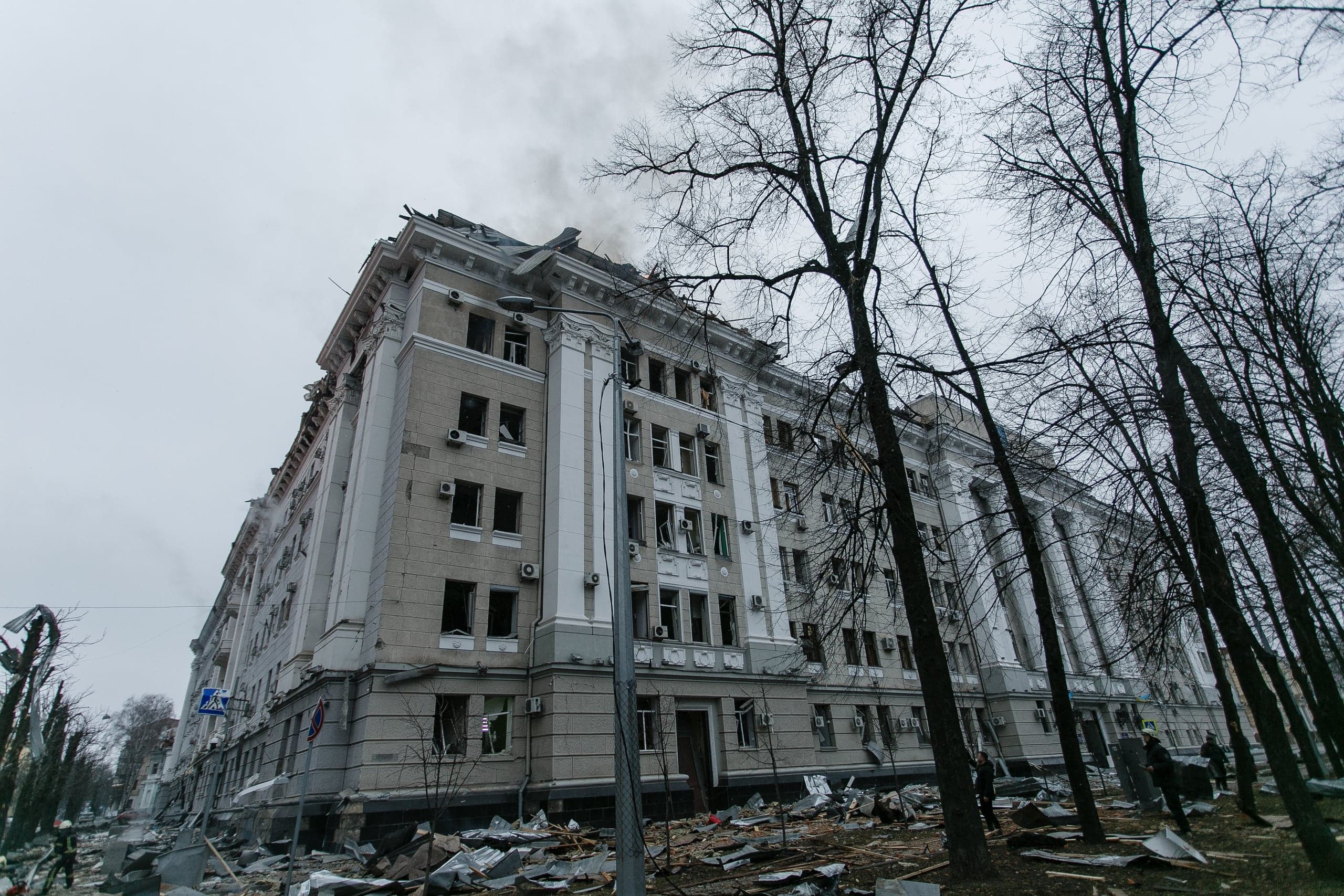 Поврежденное, частично разрушенное здание с дымящейся крышей и выбитыми окнами, вокруг обломки. Перед фасадом голые деревья.