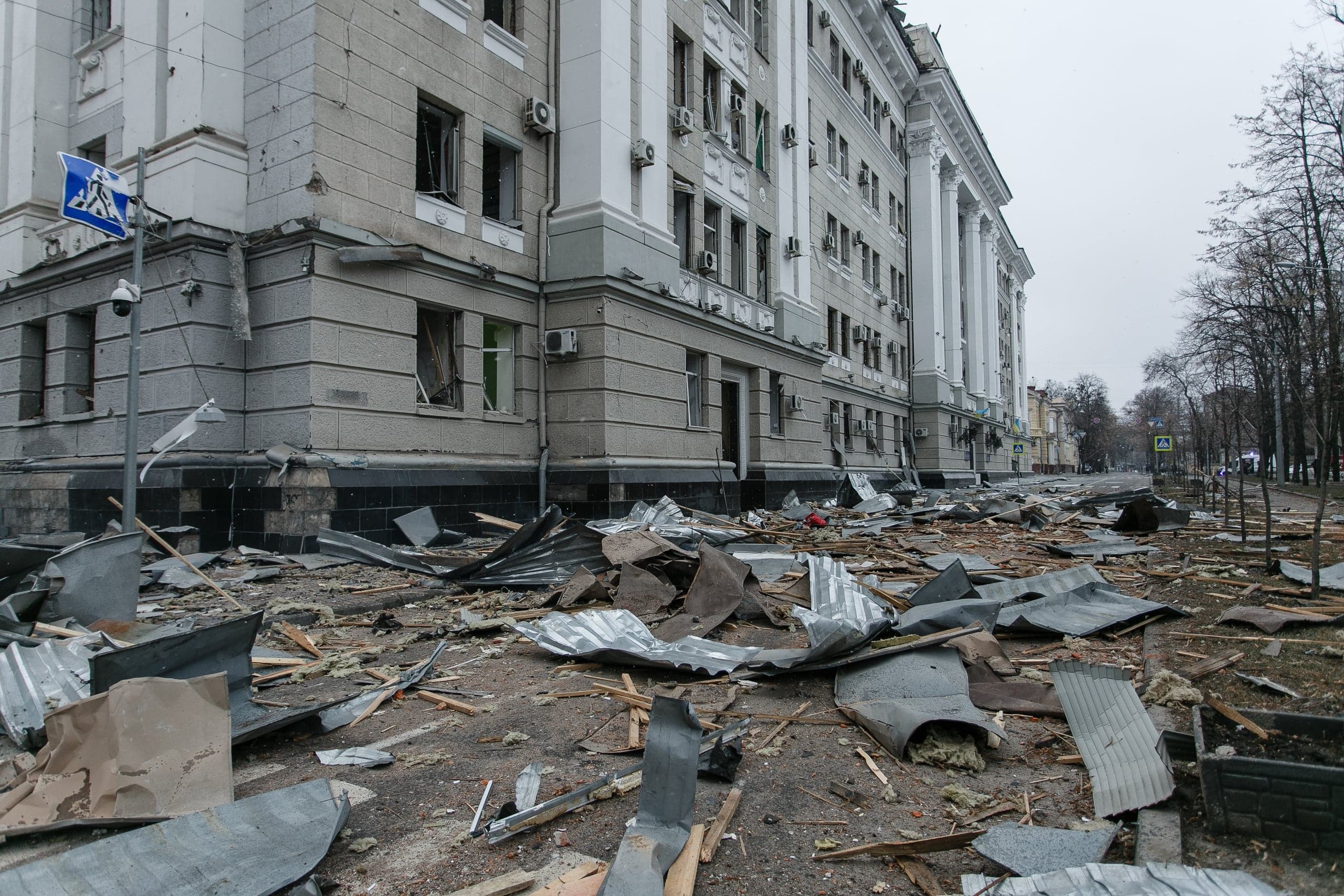 Разрушенное здание с выбитыми окнами. Улица перед фасадом усеяна обломками, скрученным металлом и строительным мусором.