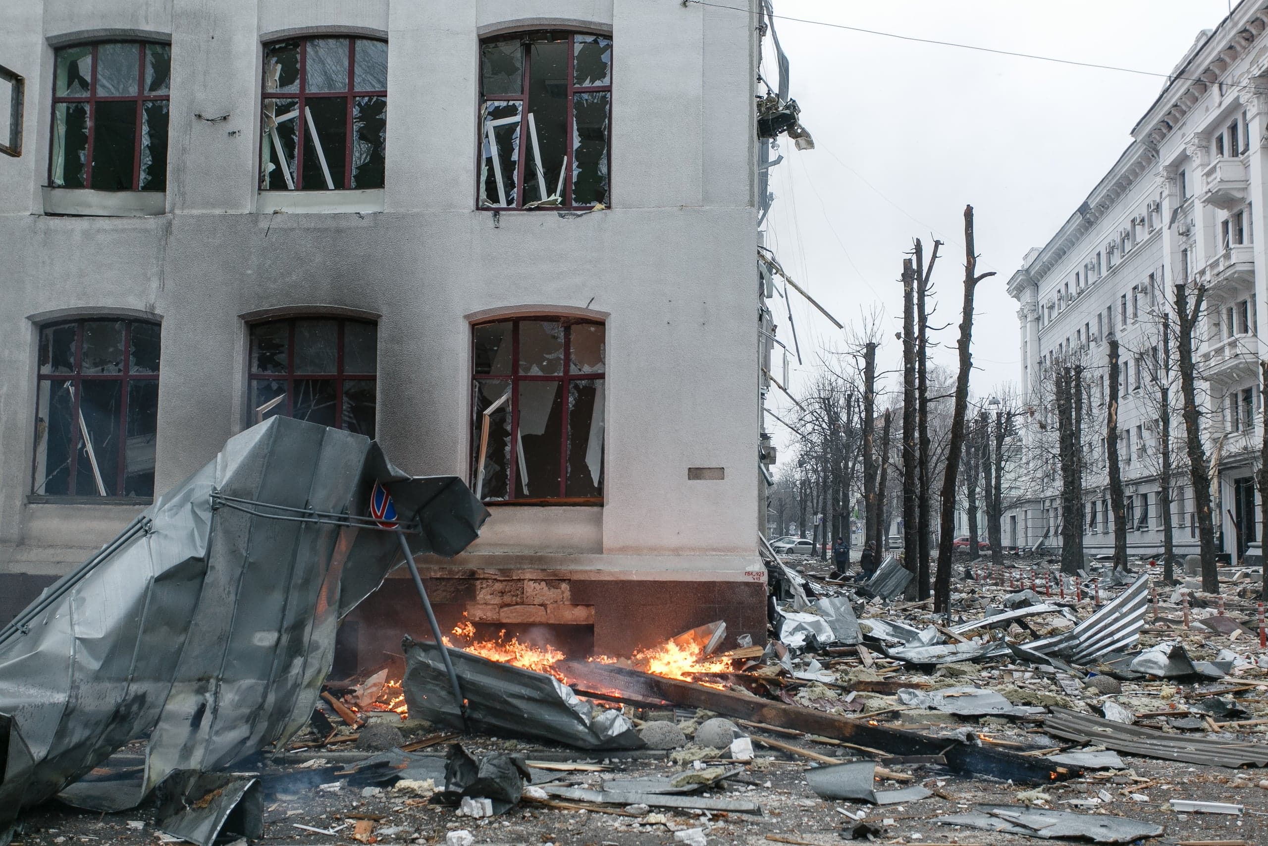 Поврежденное здание с выбитыми окнами и копотью, перед которым горит огонь среди обломков на улице. Справа видны спиленные стволы деревьев и уцелевший дом.