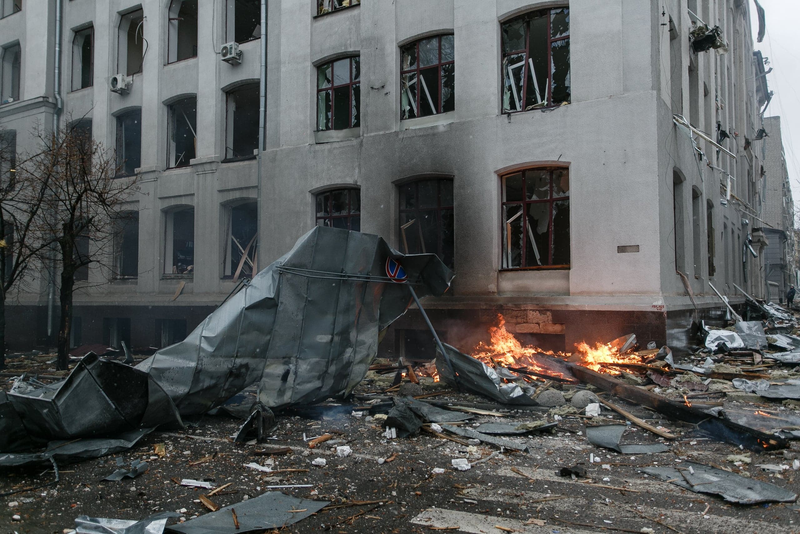 Разрушенное здание с выбитыми окнами, перед которым горит огонь и лежат обломки, включая смятую металлическую конструкцию.