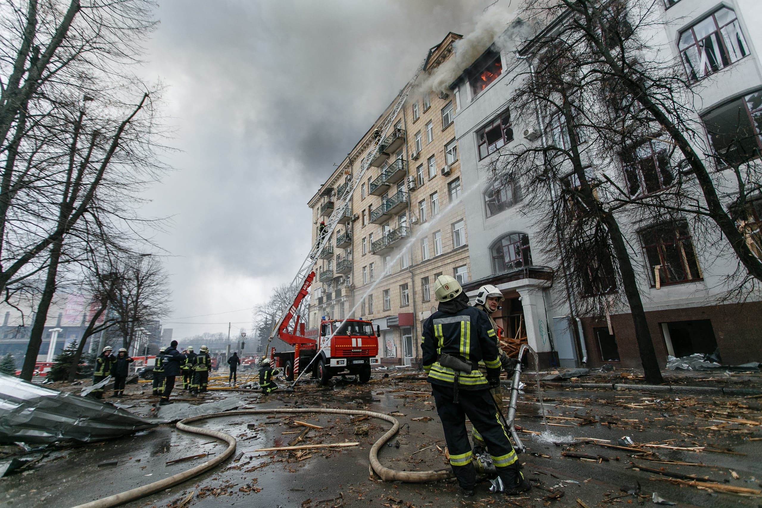 Пожарные тушат водой горящее многоэтажное здание с помощью автолестницы. Улица завалена обломками, из окон идет густой дым.
