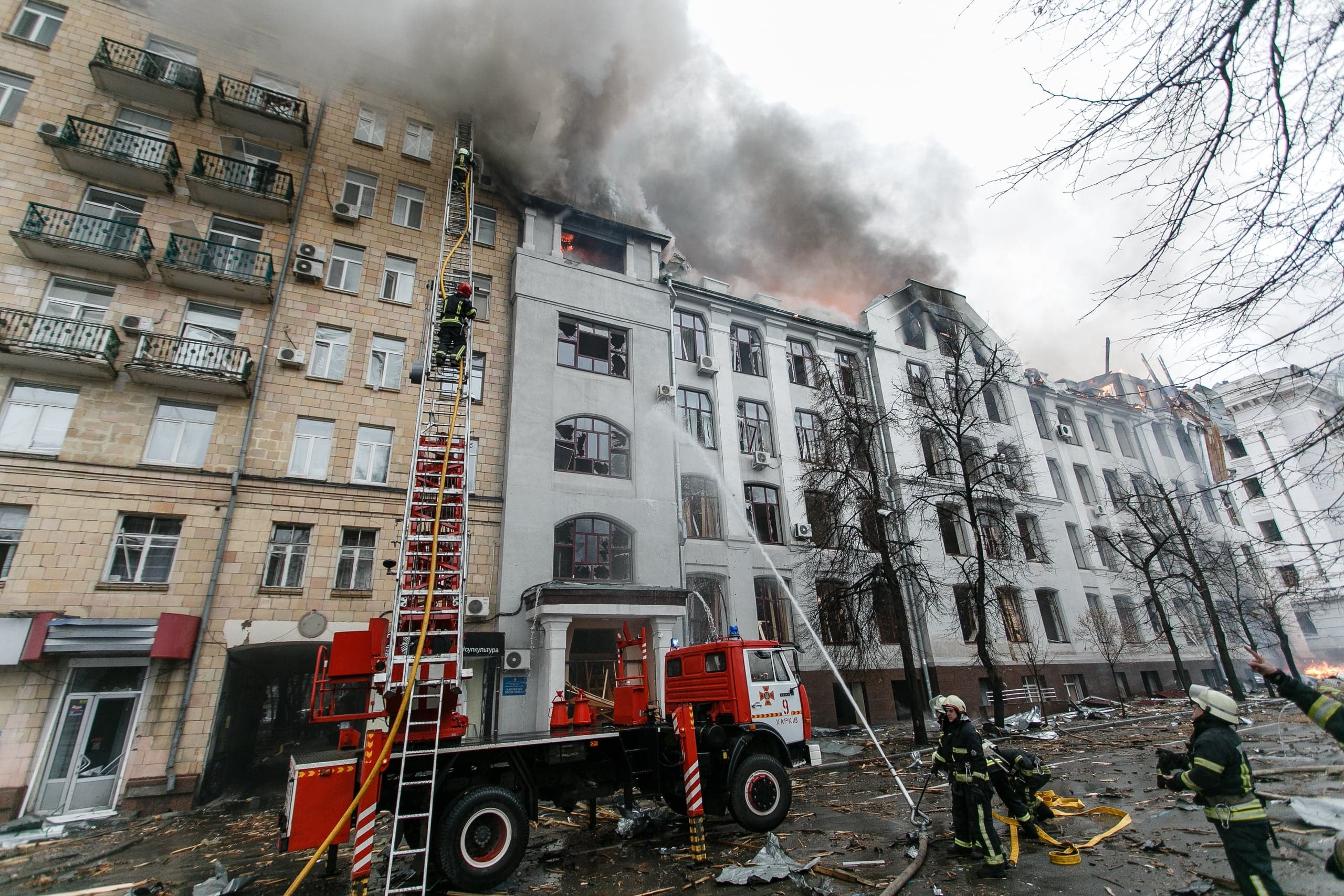 Тушение сильного пожара в многоэтажном здании. Густой дым, пламя на верхних этажах, пожарная машина с выдвижной лестницей и спасатели.