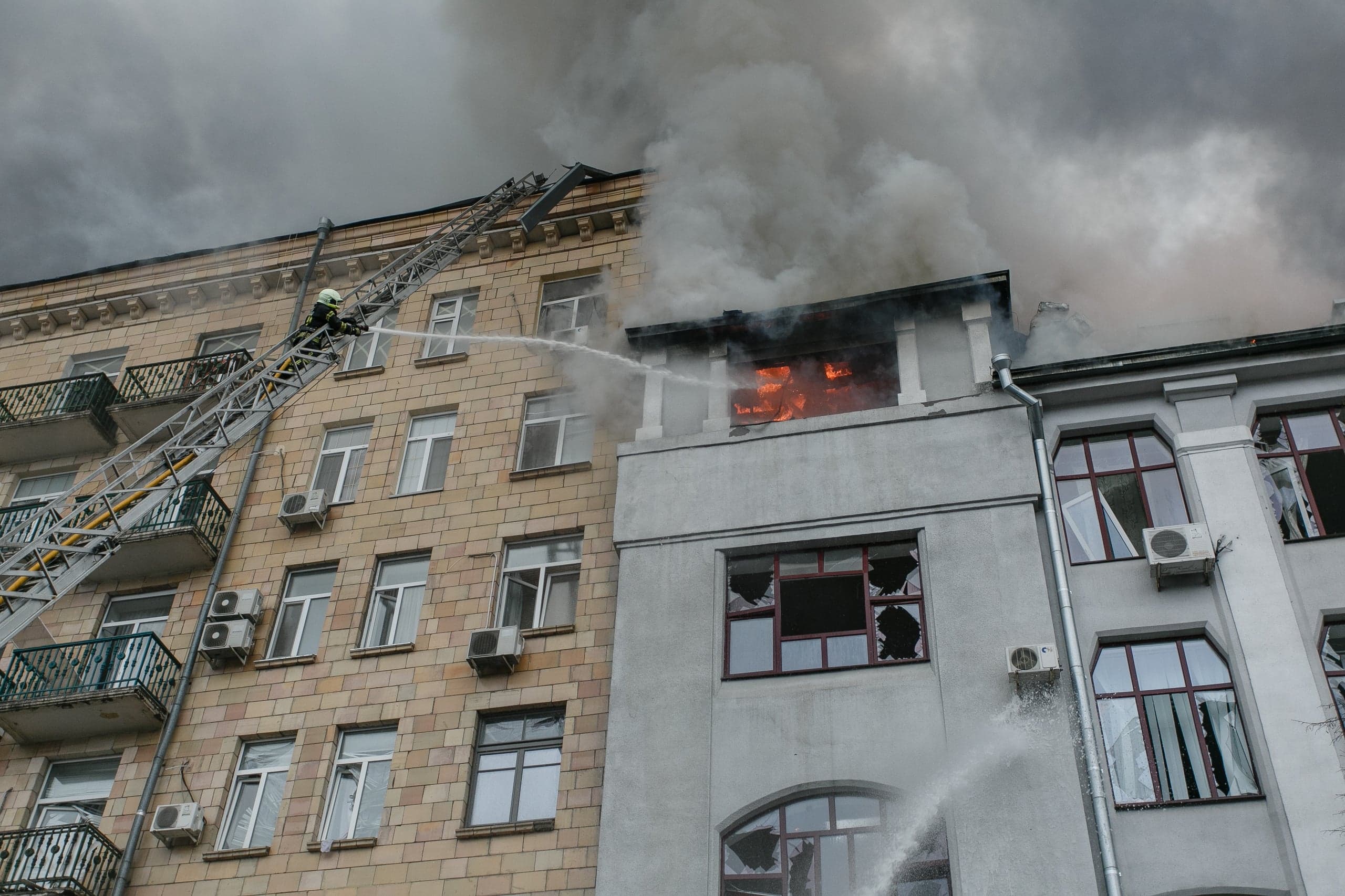 Пожарный на выдвижной лестнице тушит сильный огонь и дым в окне верхнего этажа здания.