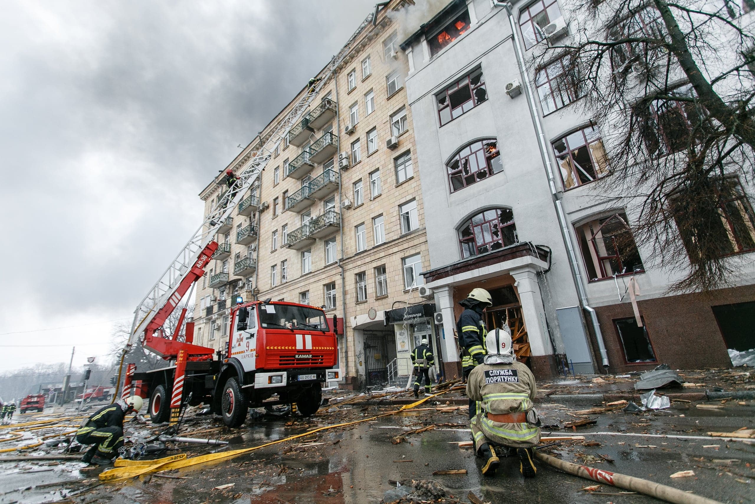 Пожарные и спецтехника на улице после обстрела. Огонь виден на верхних этажах поврежденного здания. Красная пожарная машина с выдвижной лестницей стоит среди обломков. На форме спасателя "СЛУЖБА ПОРЯТУНКУ".