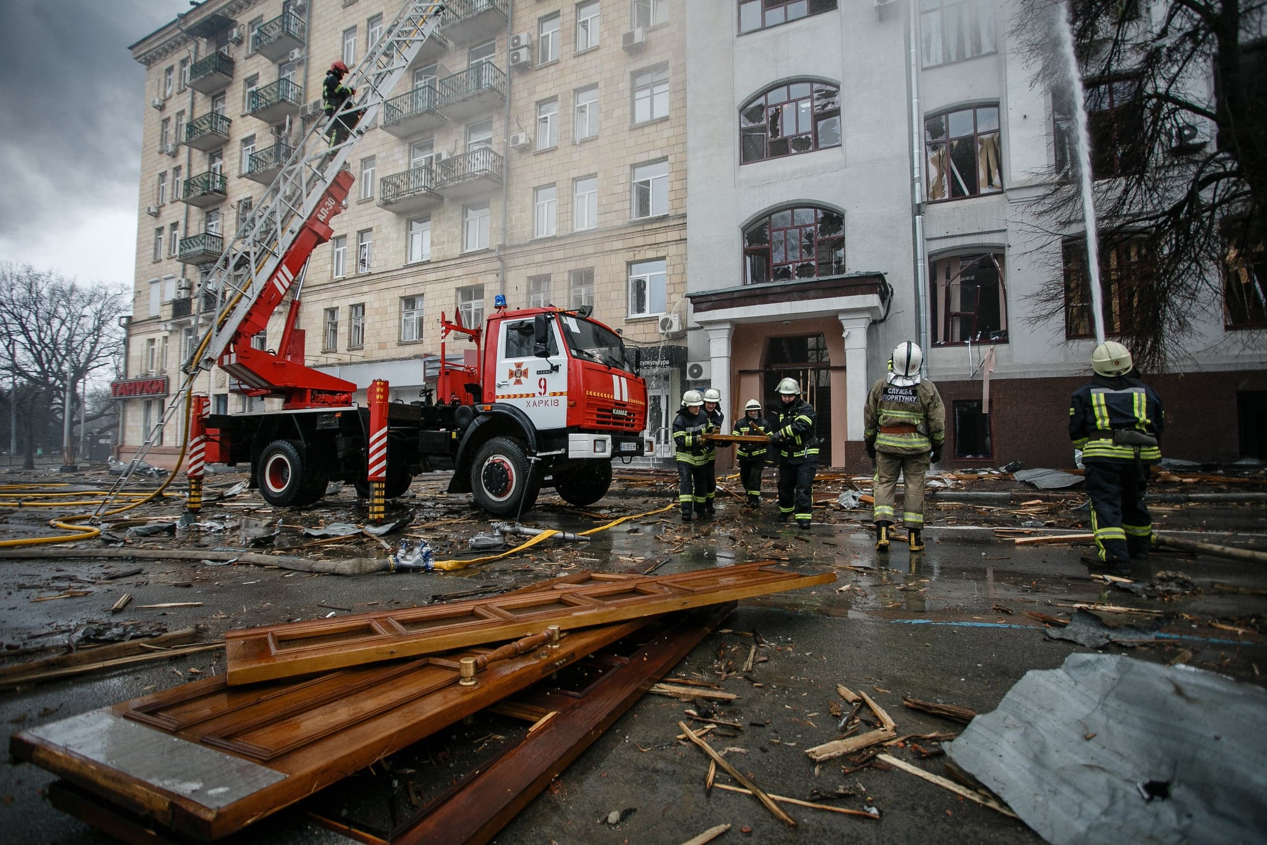 Спасатели ГСЧС с пожарной машиной и выдвижной лестницей работают на месте разрушенного здания. Улица завалена обломками и обгоревшими досками. На машине виден текст "ХАРКІВ".