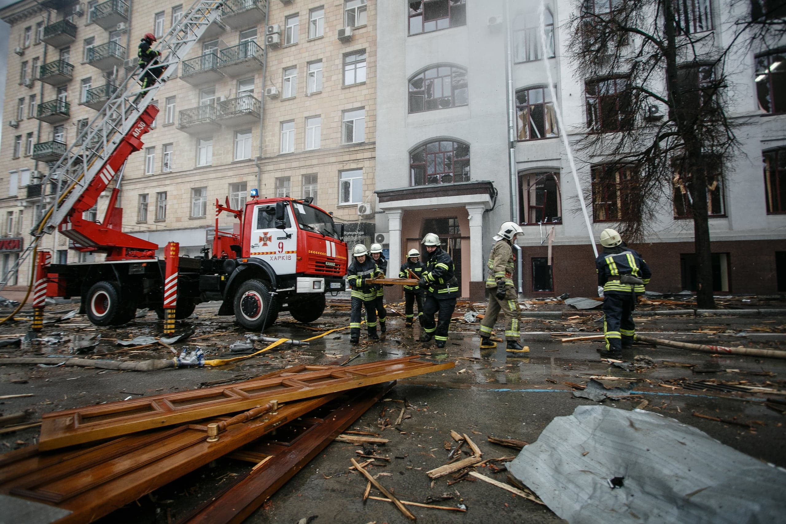 Спасатели ликвидируют последствия разрушения здания в Харькове. Пожарная машина с надписью "9 ХАРКІВ" и выдвинутой лестницей, сотрудники тушат поврежденный фасад и убирают обломки на улице.