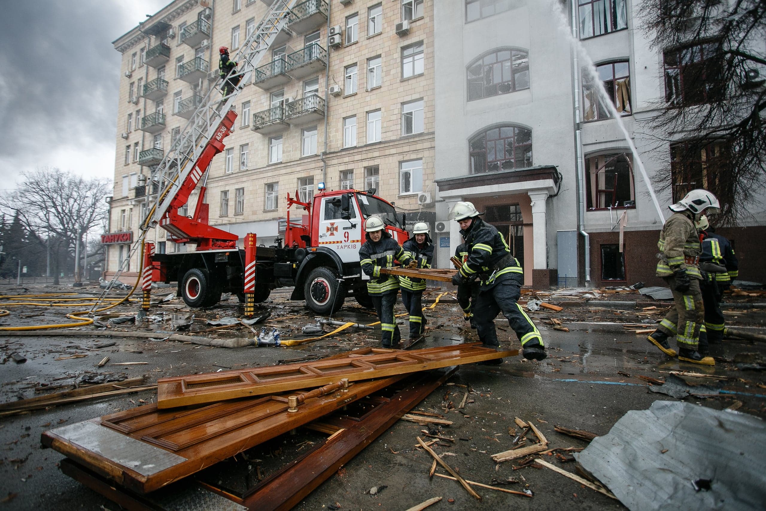 Пожарные разбирают завалы и поливают водой поврежденное здание. На улице обломки и сломанные двери. На пожарной машине видна надпись "ХАРКІВ".