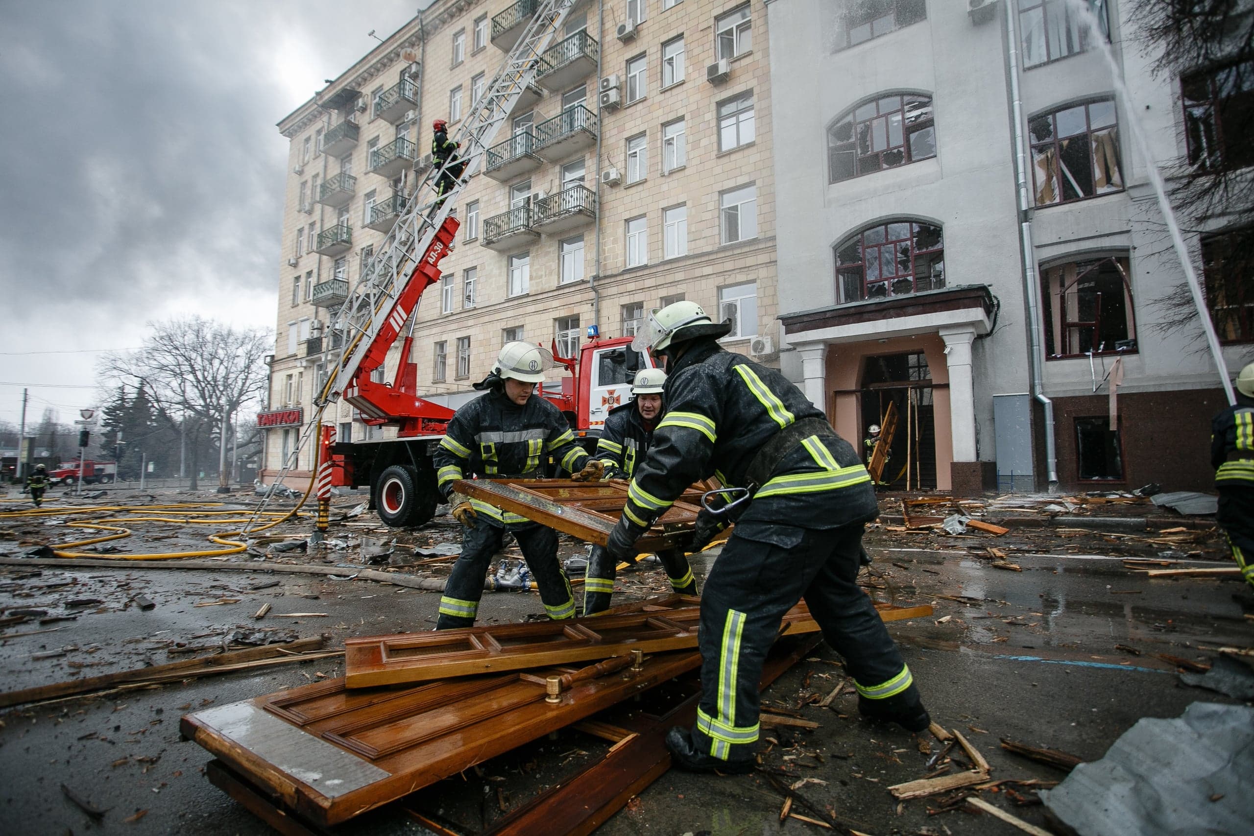 Три спасателя разбирают деревянные обломки перед зданием с разбитыми окнами. На заднем плане пожарная автолестница.