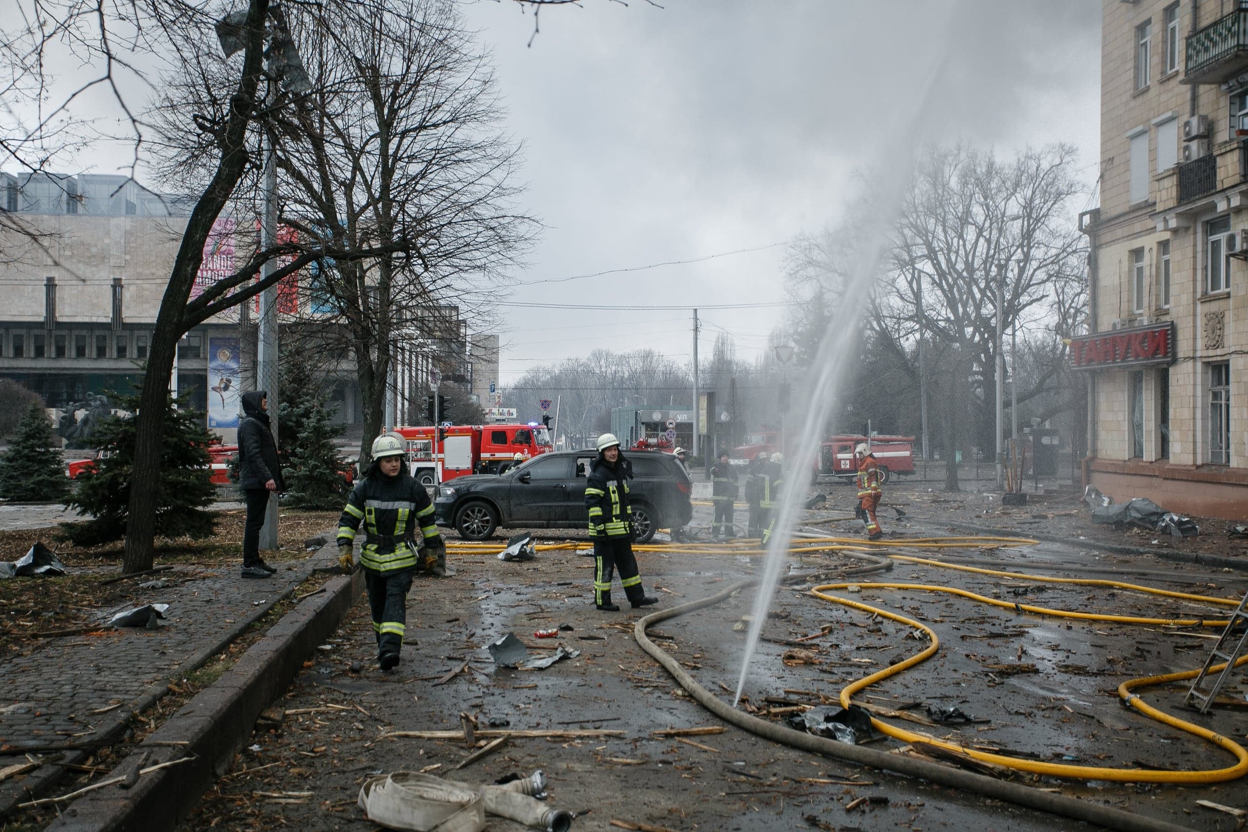 Пожарные работают на городской улице, покрытой обломками и шлангами, после происшествия. Видна мощная струя воды из шланга. Красные пожарные машины на заднем плане.