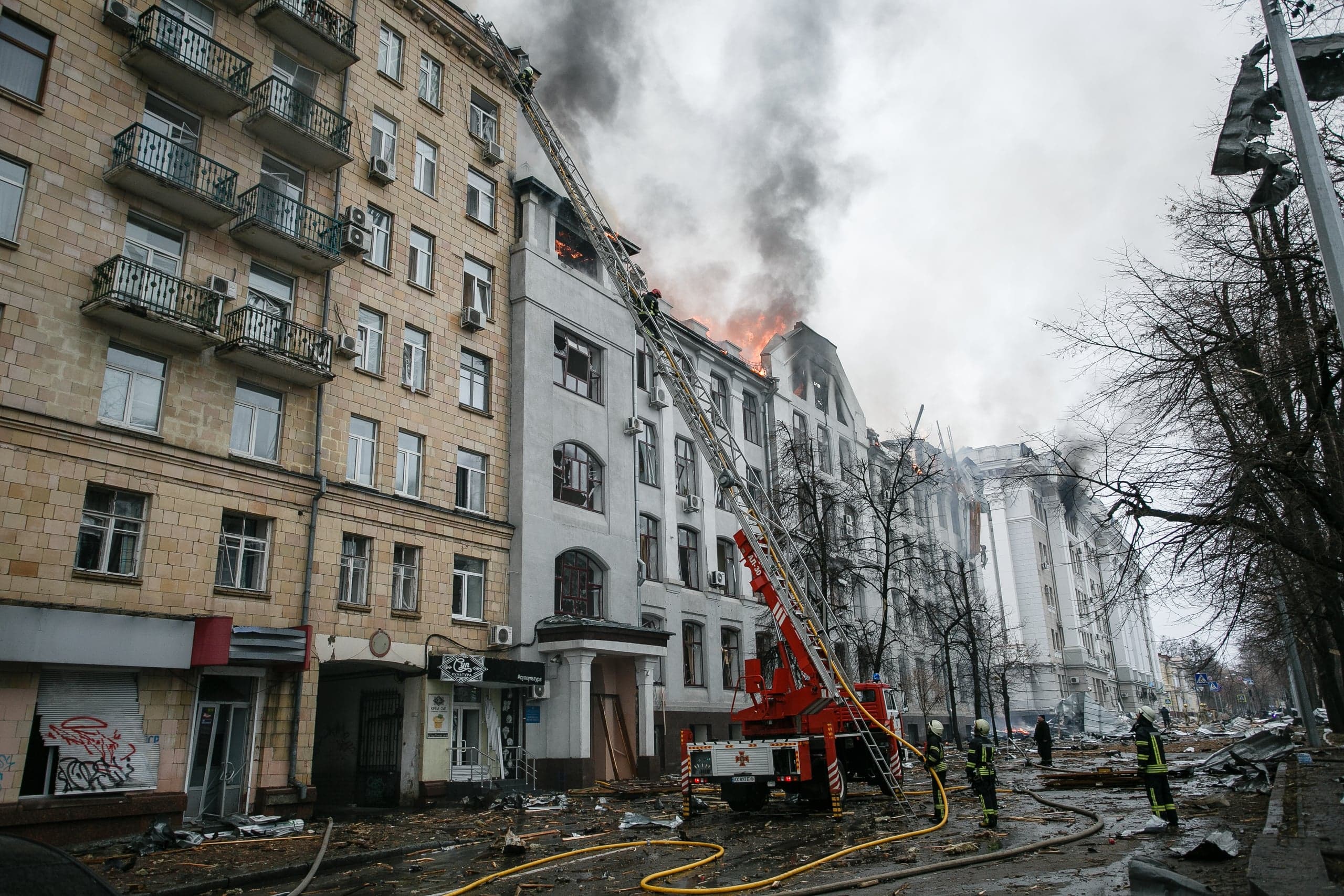 Пожарные тушат горящее многоэтажное здание с помощью автолестницы; улица усыпана обломками и дымом.