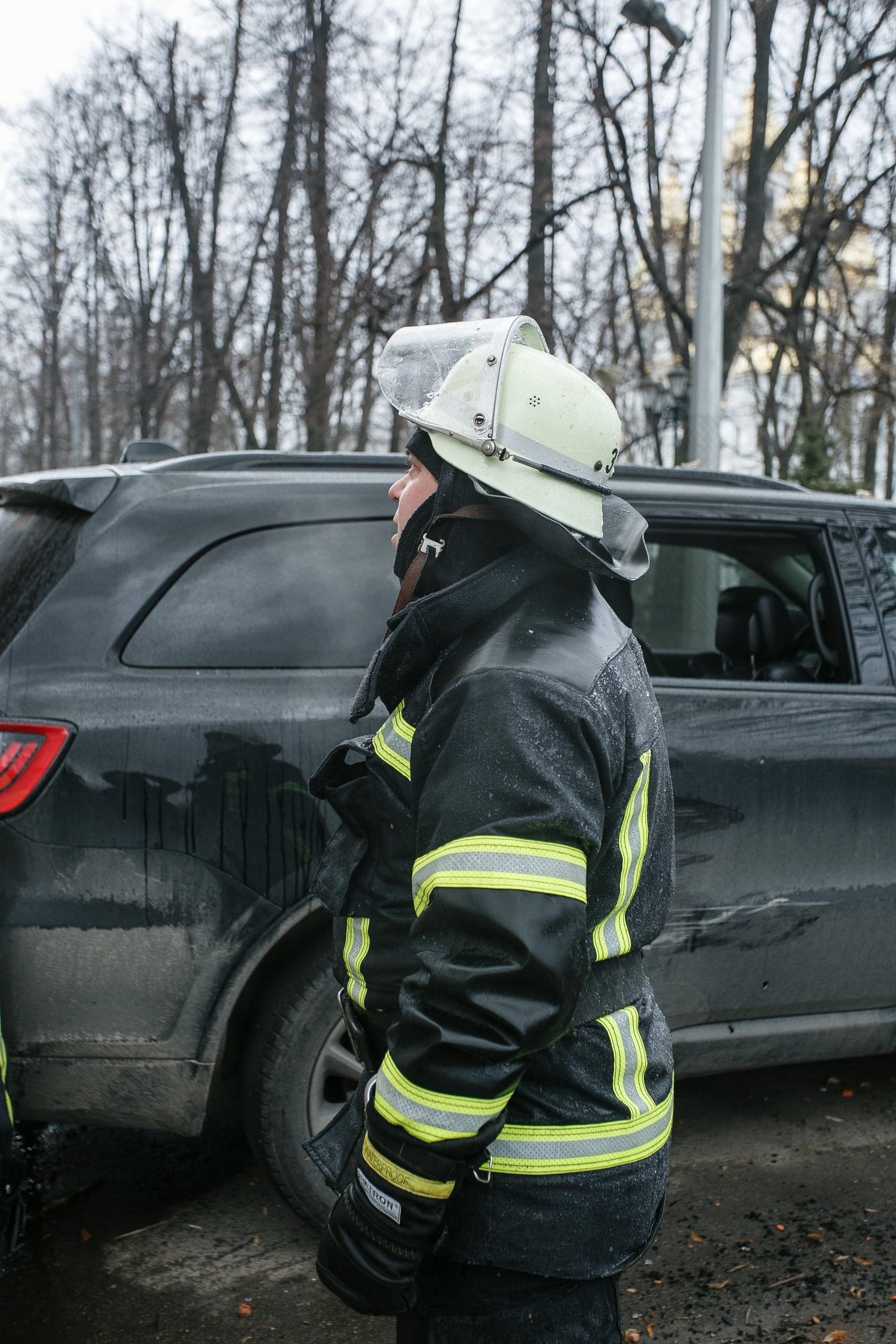 Пожарный в защитной форме и шлеме стоит боком на улице на фоне черного внедорожника и деревьев.