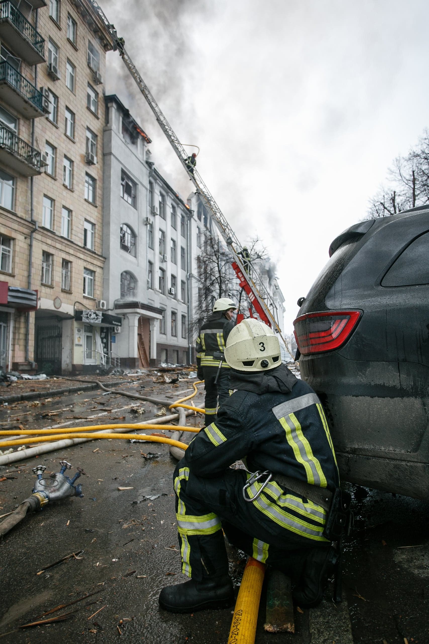 Пожарный сидит на мокрой улице среди желтых шлангов и обломков, на фоне горящего многоэтажного здания. Спасатели по выдвижной лестнице тушат огонь на верхних этажах.