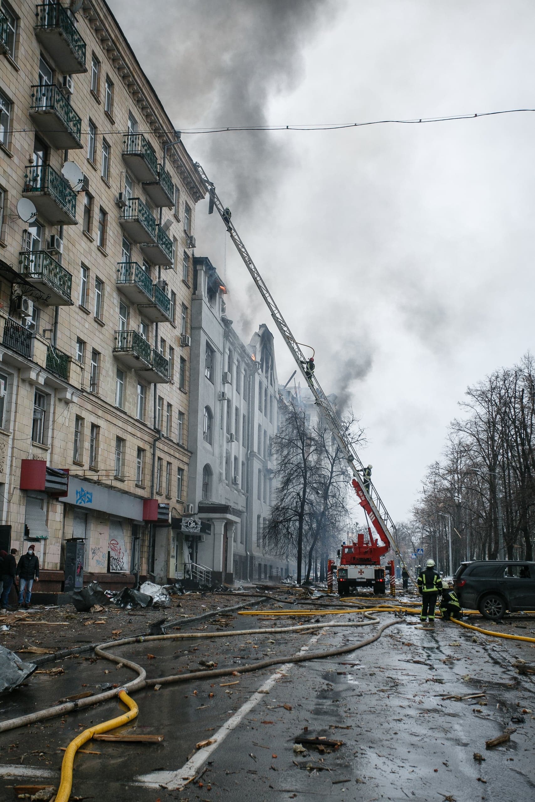Пожарные тушат огонь на верхних этажах поврежденного городского здания с помощью автолестницы. Густой дым и обломки на мокрой улице.