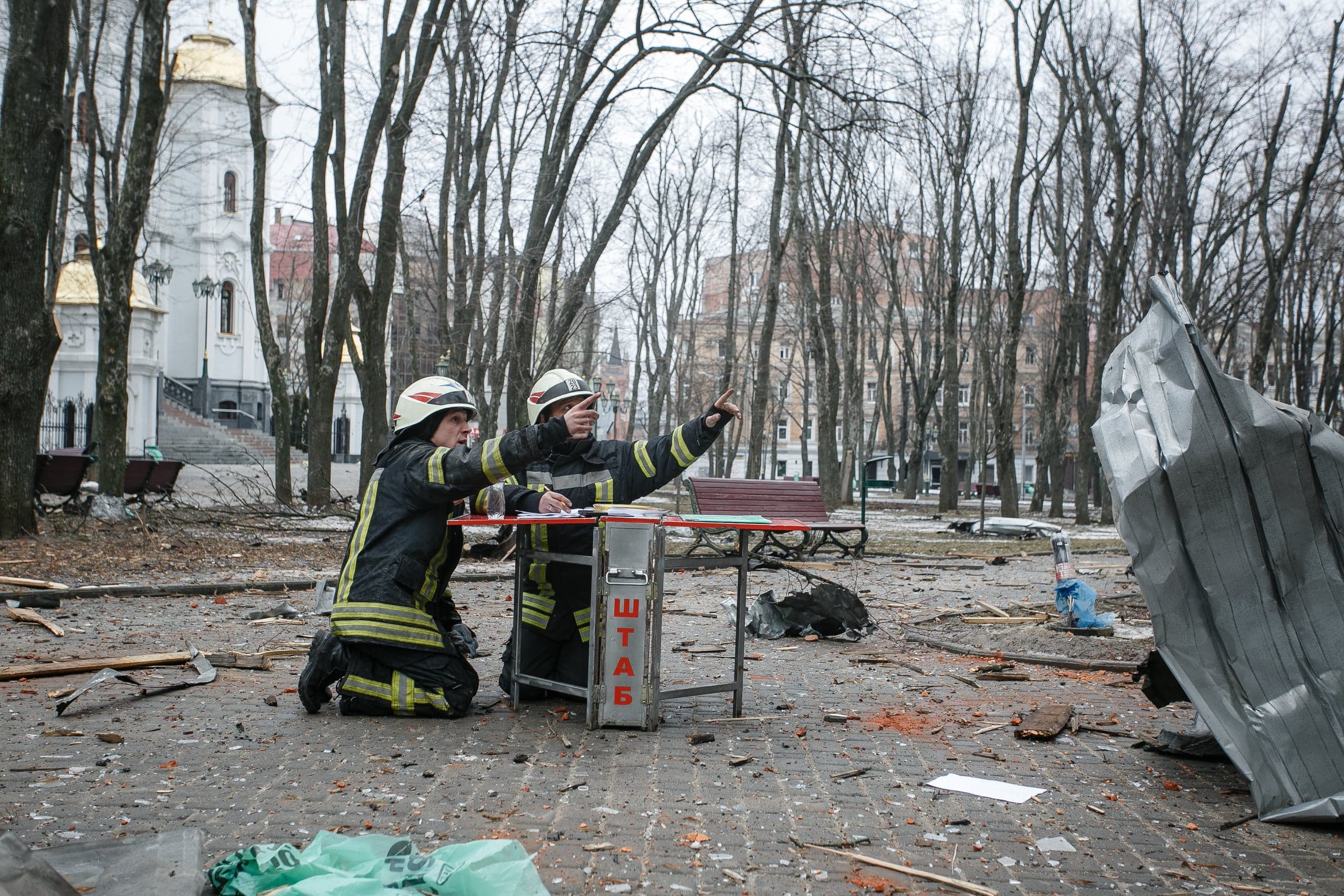 Два спасателя осматривают парк, усыпанный обломками после удара, стоя у стола с надписью "ШТАБ". На заднем плане виден собор с золотыми куполами.