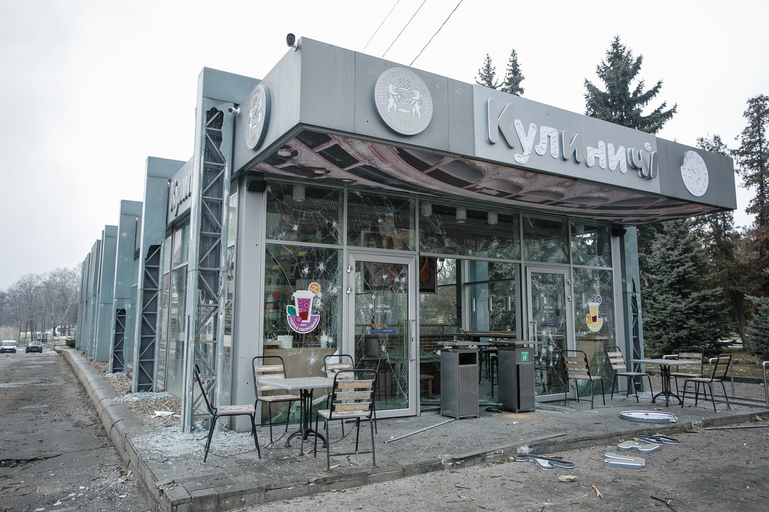 Фасад магазина "Кулиничі" с витринами, пробитыми осколками. На тротуаре осколки стекла и упавшие части вывески.