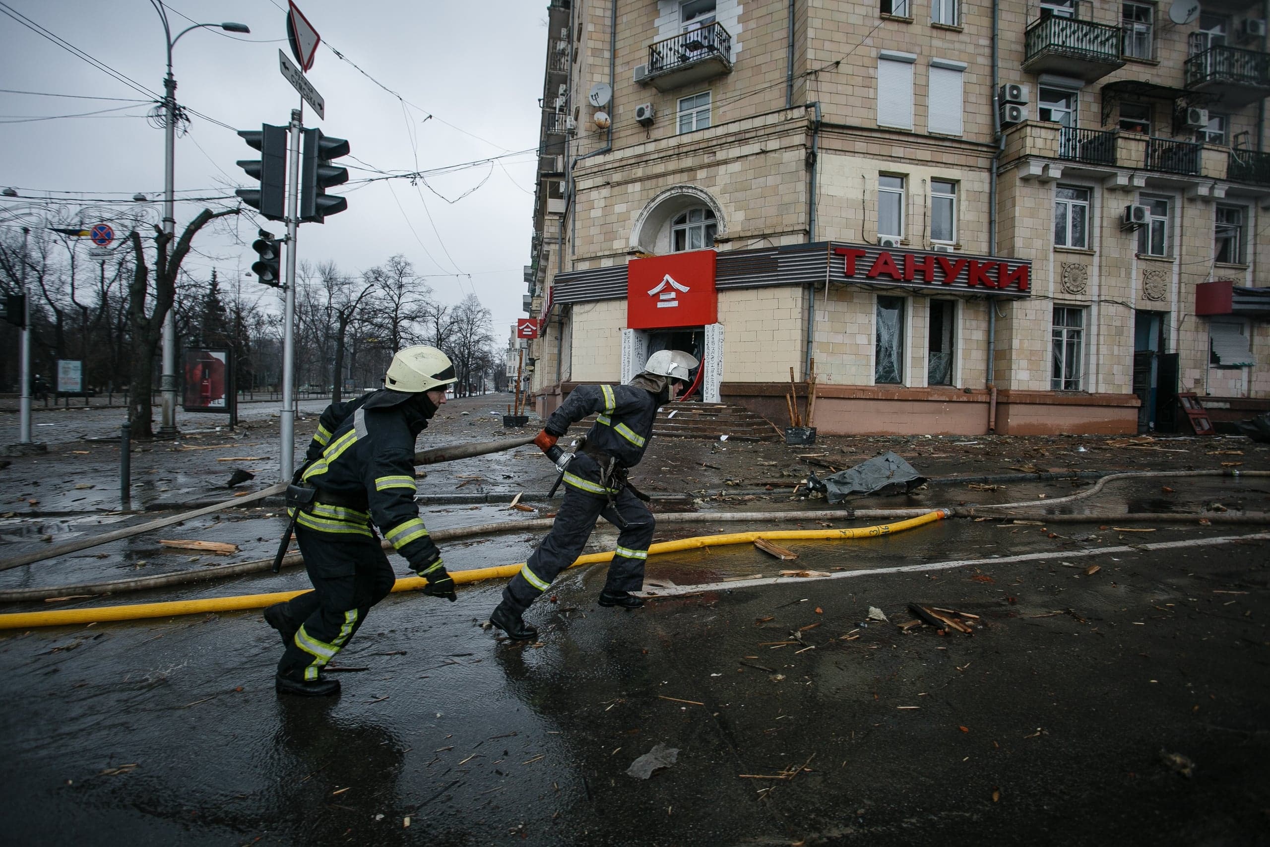 Два пожарных бегут по мокрой, усыпанной обломками улице мимо поврежденного здания с вывеской "ТАНУКИ".