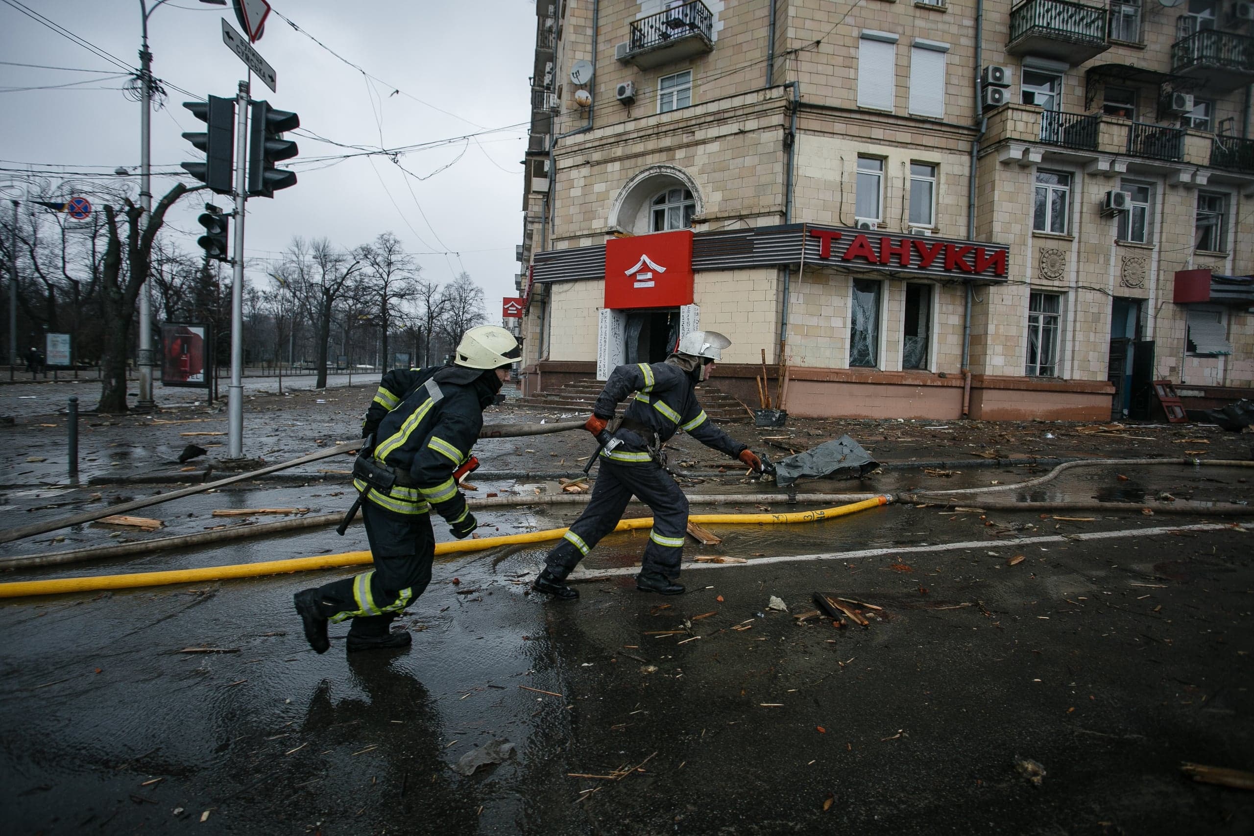 Два пожарных спешат по заваленной обломками улице возле поврежденного здания с вывеской "ТАЙНКИ".