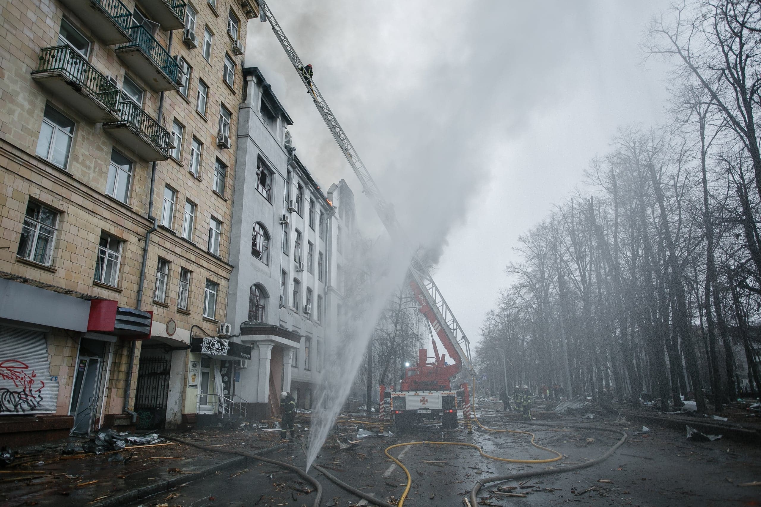 Пожарные тушат горящее здание с помощью автолестницы и брандспойтов. Из окон идет густой дым. На улице много обломков.