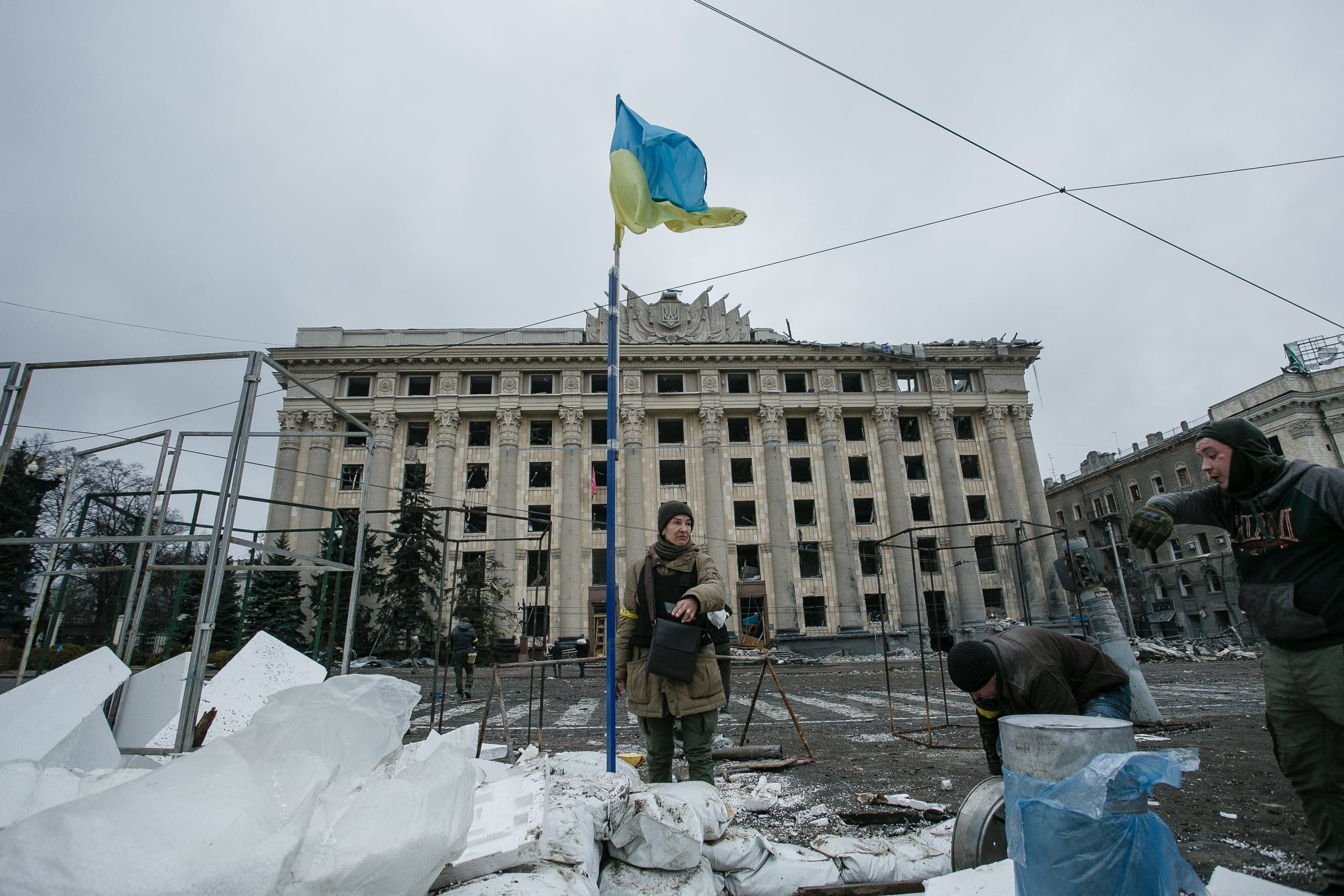 Разрушенное здание Харьковской ОГА с выбитыми окнами и поврежденной крышей. Перед зданием — баррикада из мешков с песком, украинский флаг и несколько военных или гражданских лиц. Пасмурная погода.