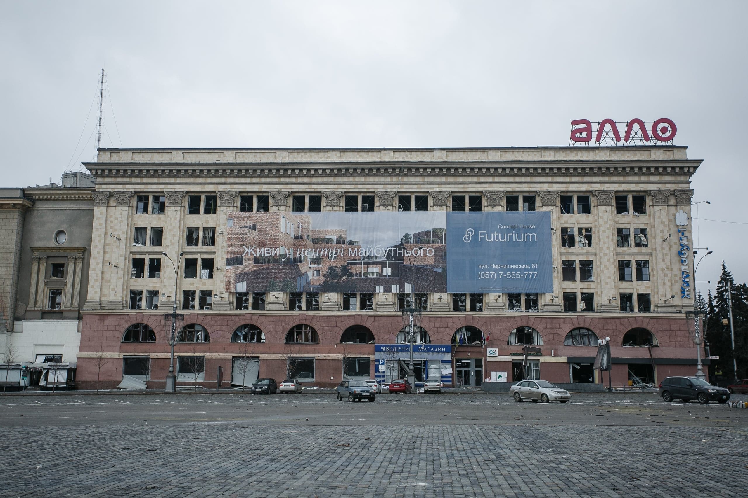 Здание с классическим фасадом, поврежденное обстрелом, с разбитыми окнами. На крыше красная вывеска "АЛЛО", на фасаде рекламный баннер с текстом "ЖИВИ У ЦЕНТРІ МАЙБУТНЬОГО".