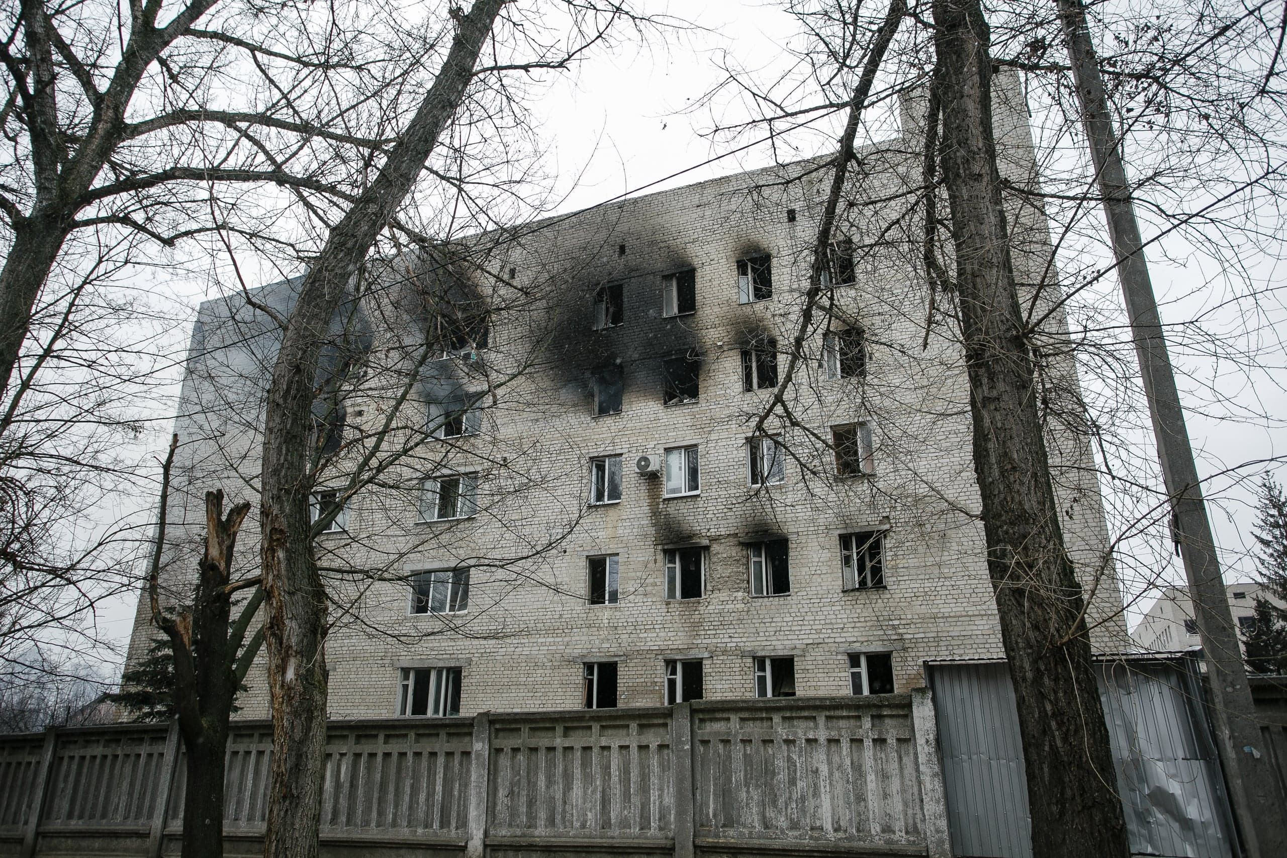 Поврежденное, закопченное огнем пятиэтажное здание из белого кирпича, вид сквозь голые деревья.