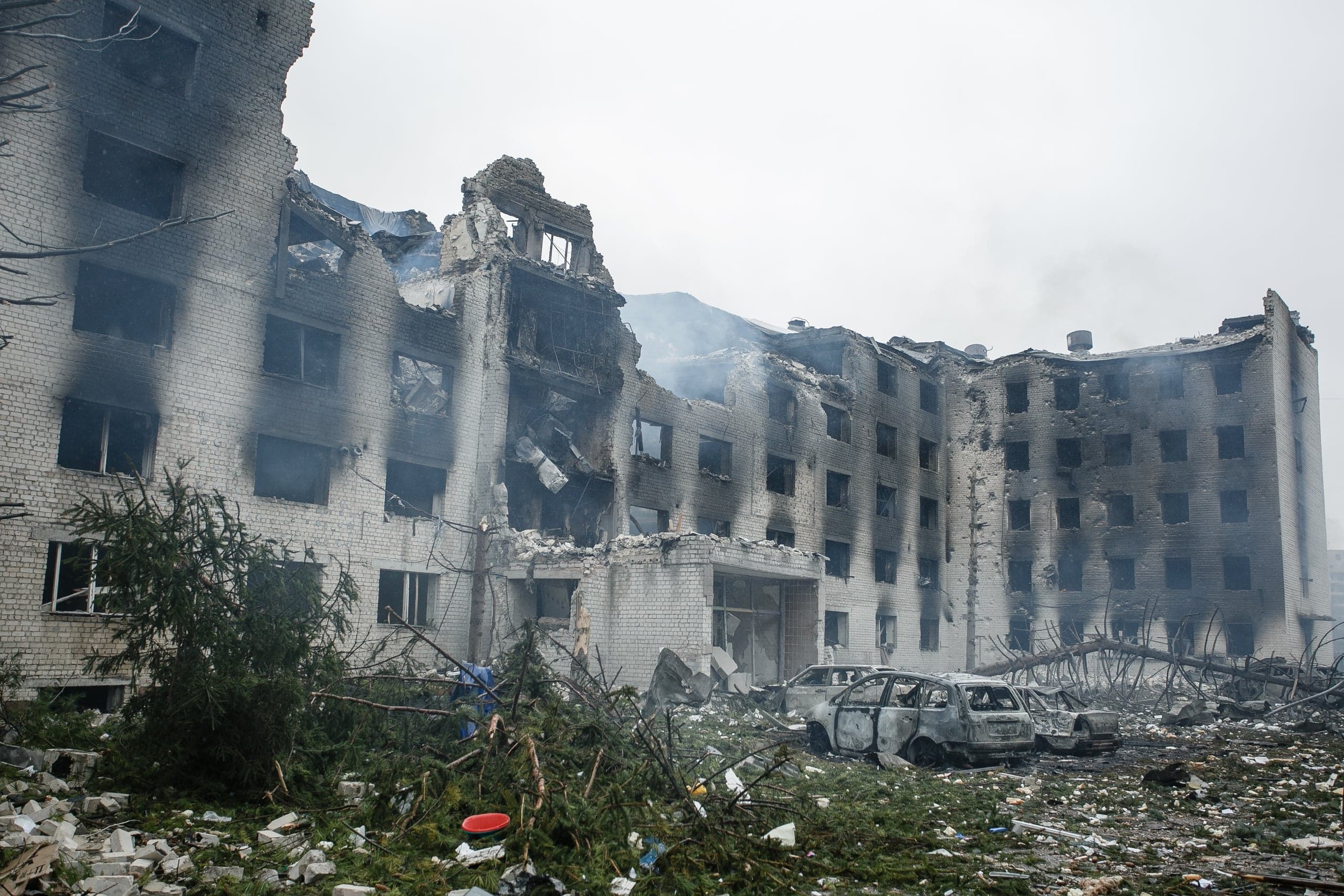 Разрушенное многоэтажное кирпичное здание с обрушенными верхними этажами и следами пожара. На земле — обломки и два сгоревших автомобиля.