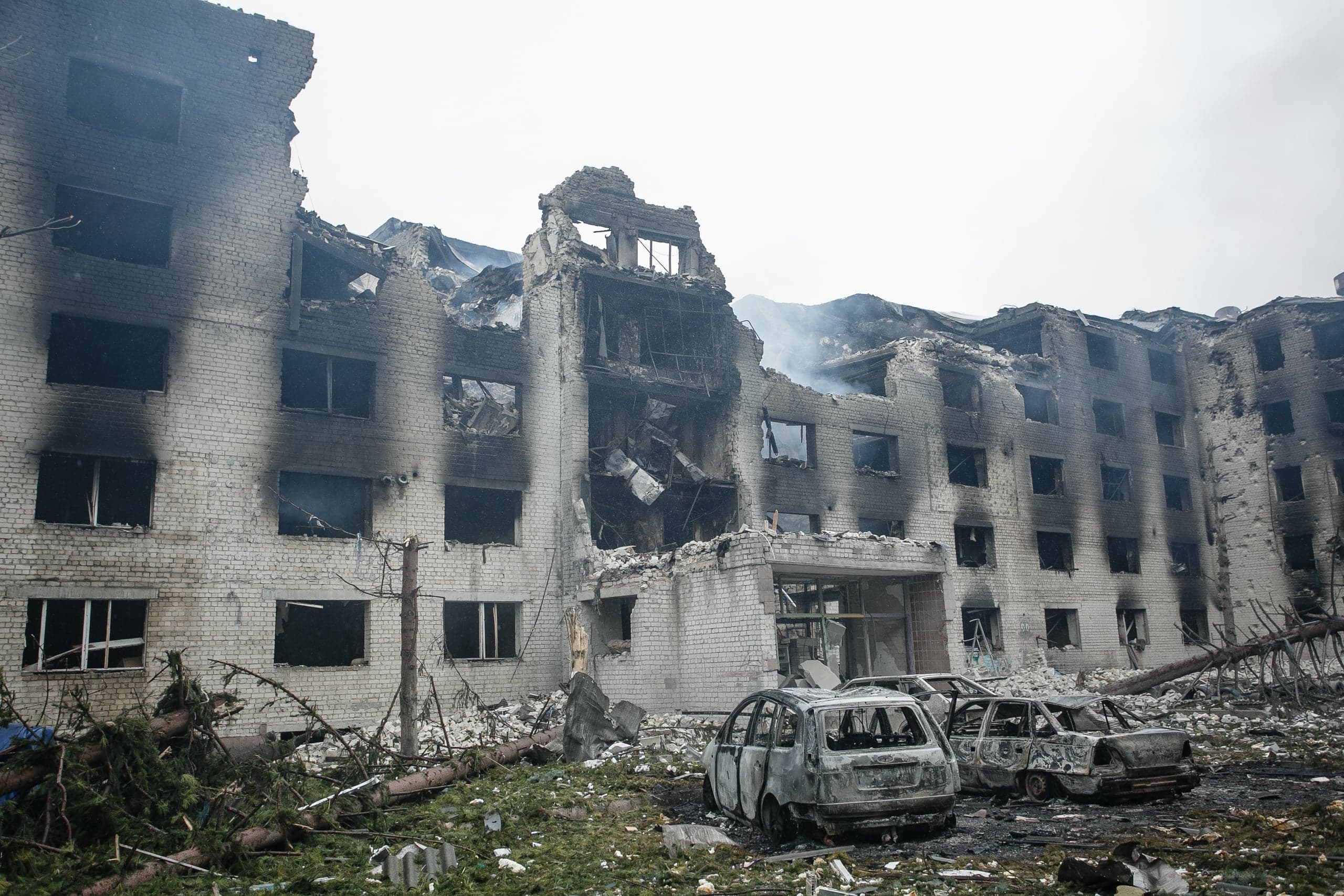 Разрушенное, обгоревшее многоэтажное здание с выбитыми окнами и частично обрушенной крышей; перед ним — три сожженных автомобиля среди обломков и поваленных деревьев.