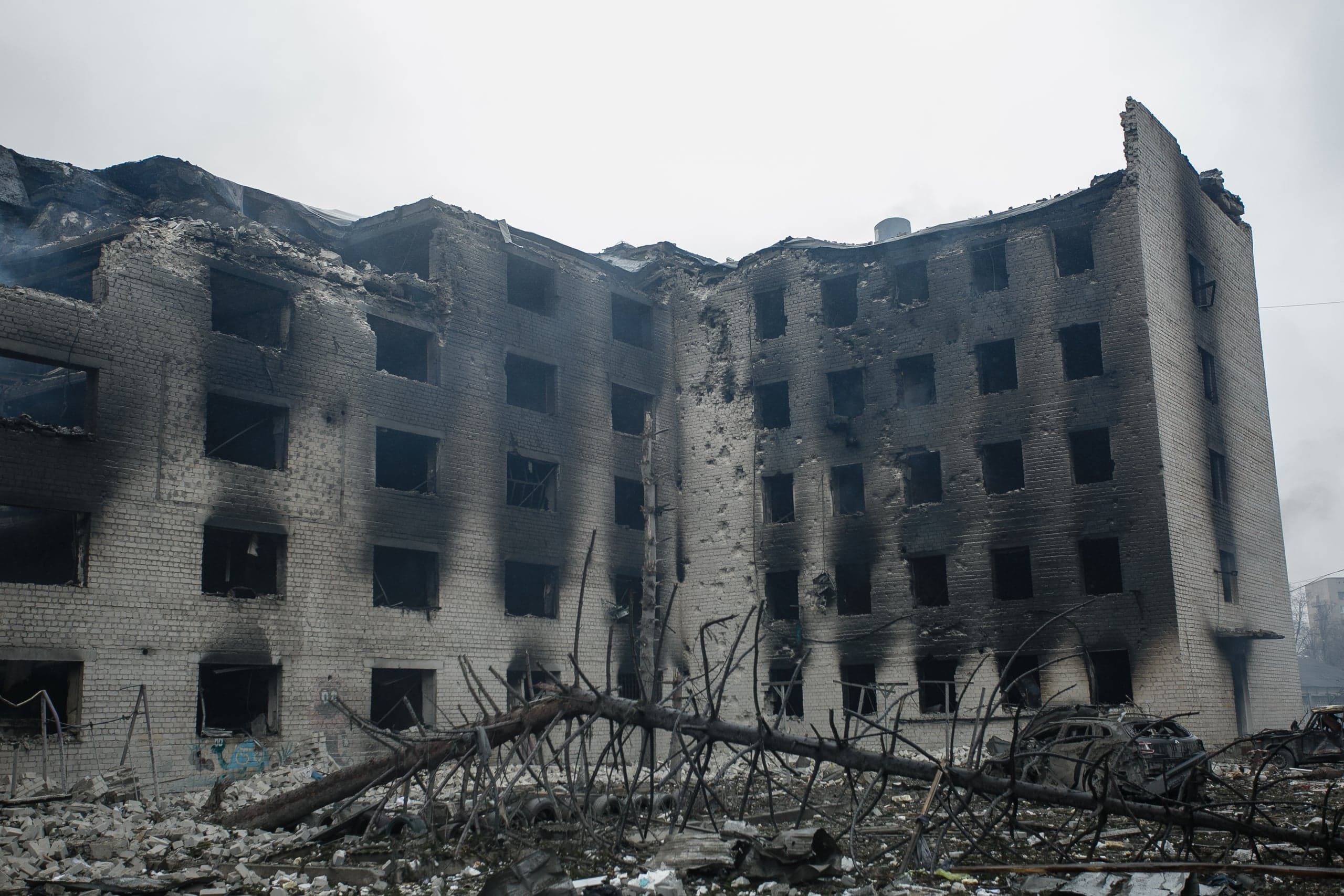 Разрушенное и обугленное многоэтажное здание с пустыми оконными проемами, окруженное строительными обломками и упавшим деревом.
