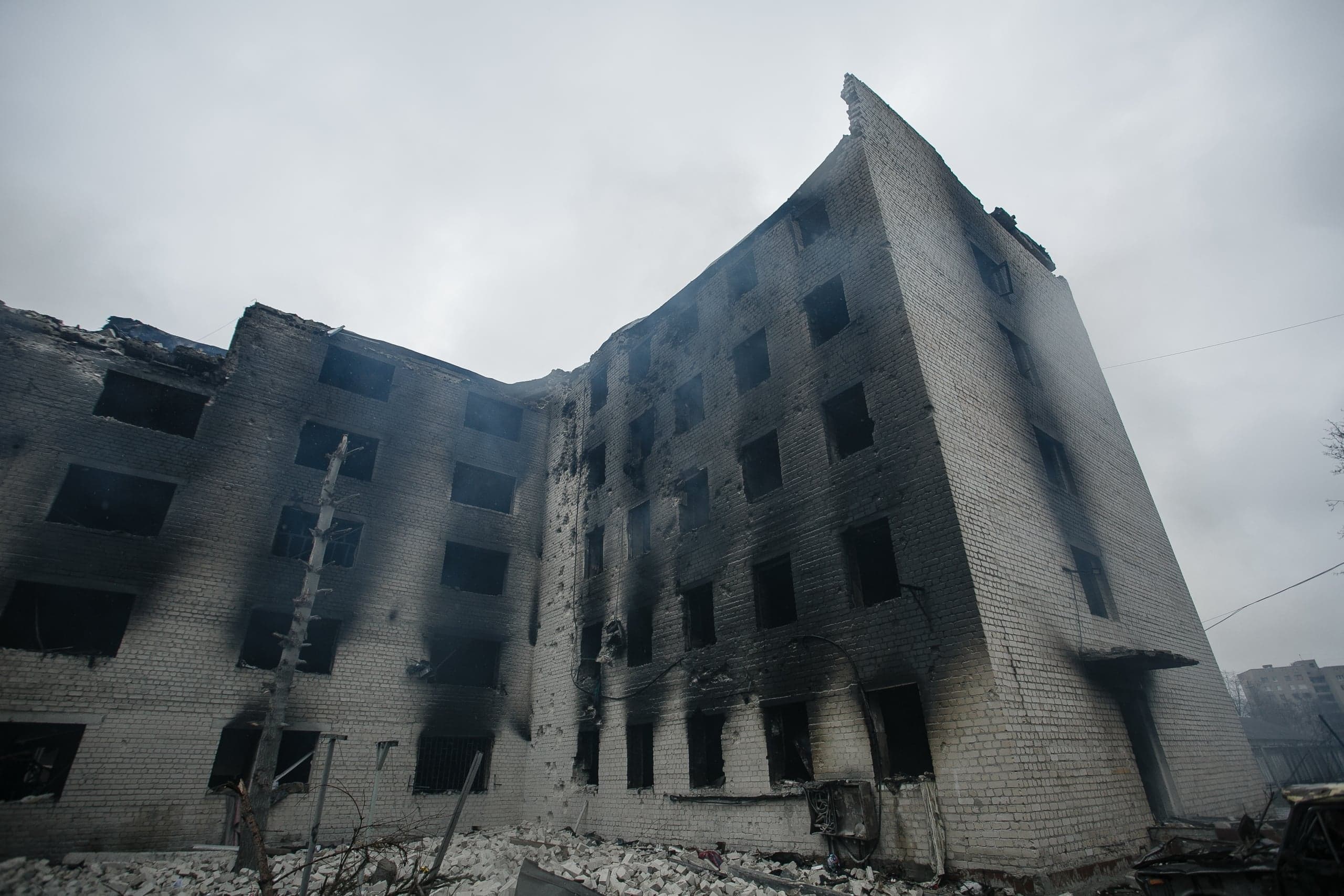 Разрушенное пятиэтажное жилое здание из белого кирпича, закопченное огнем, с выбитыми окнами и поврежденной крышей. Облачное небо.