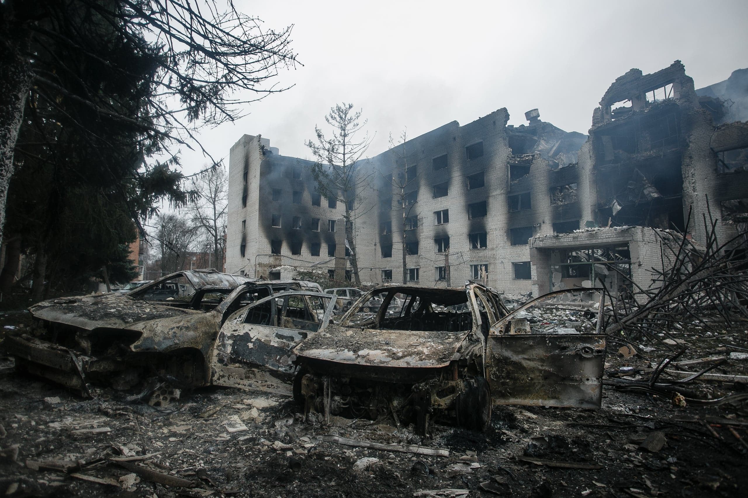 Сгоревшие остовы автомобилей перед разрушенным и обугленным многоэтажным зданием.