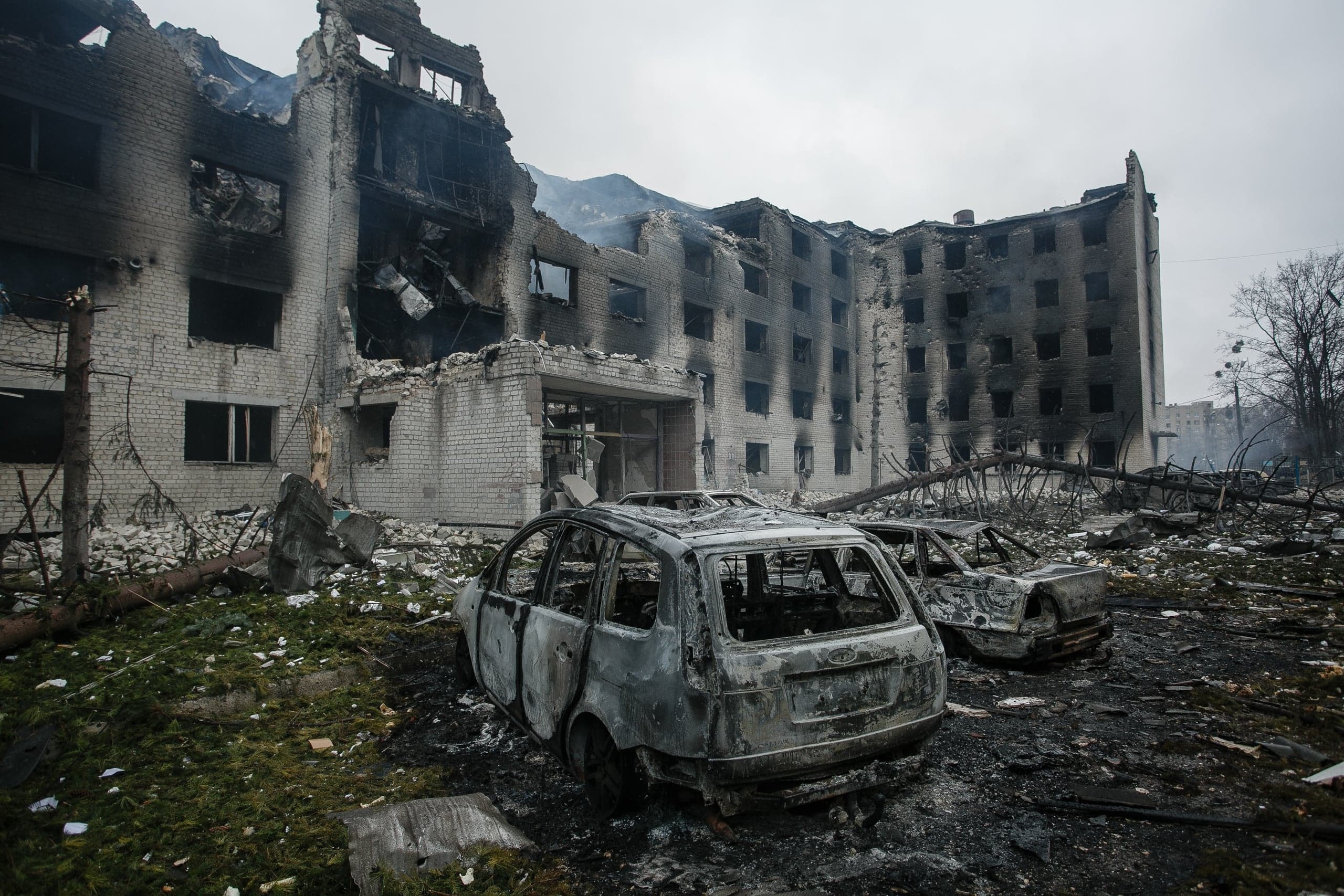 Разрушенное, обгоревшее многоэтажное здание с выбитыми окнами и частично обрушенными стенами; на переднем плане — два сгоревших остова автомобилей.