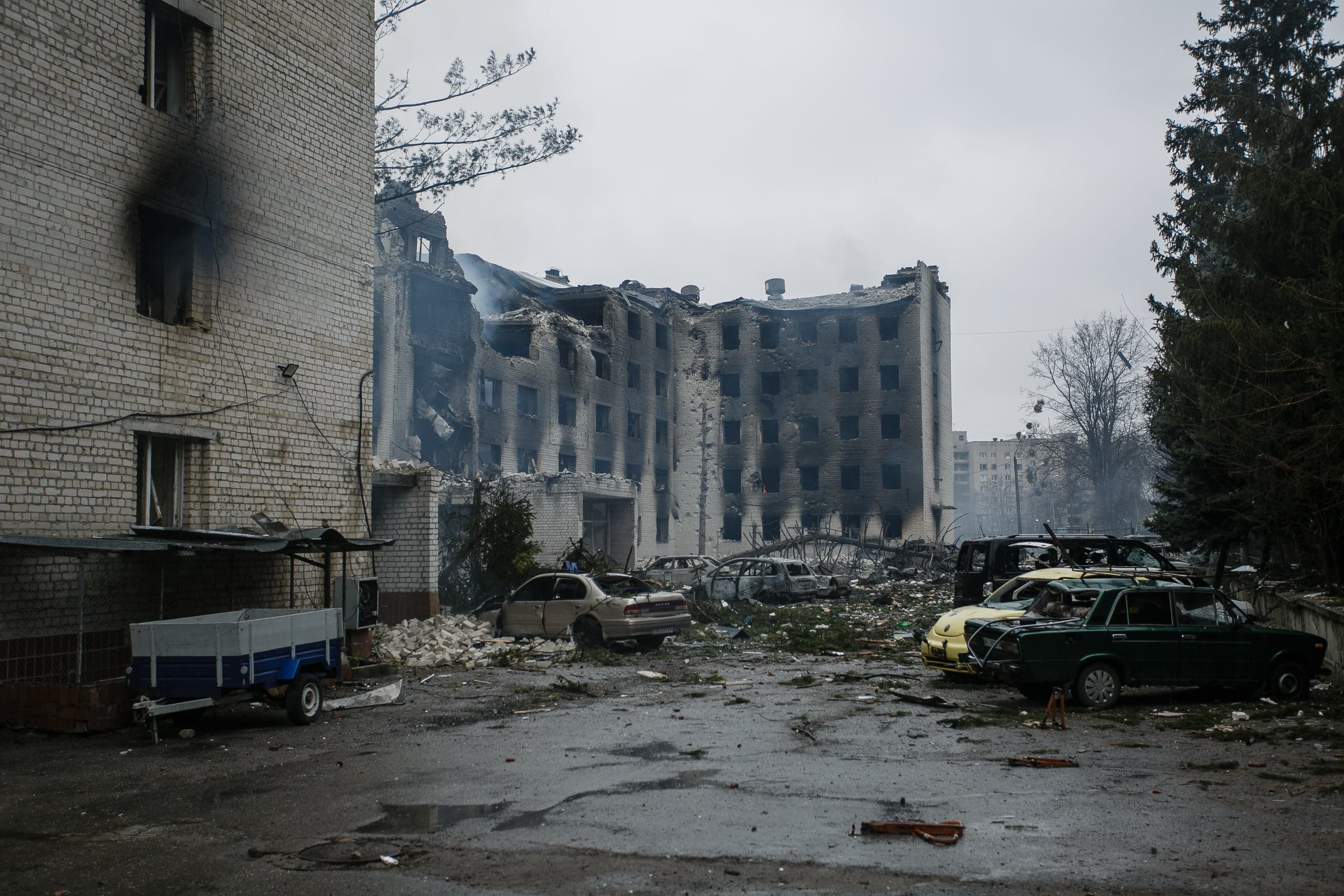 Разрушенный многоэтажный жилой дом с выгоревшей крышей и закопченными окнами, перед ним — обломки и поврежденные автомобили.