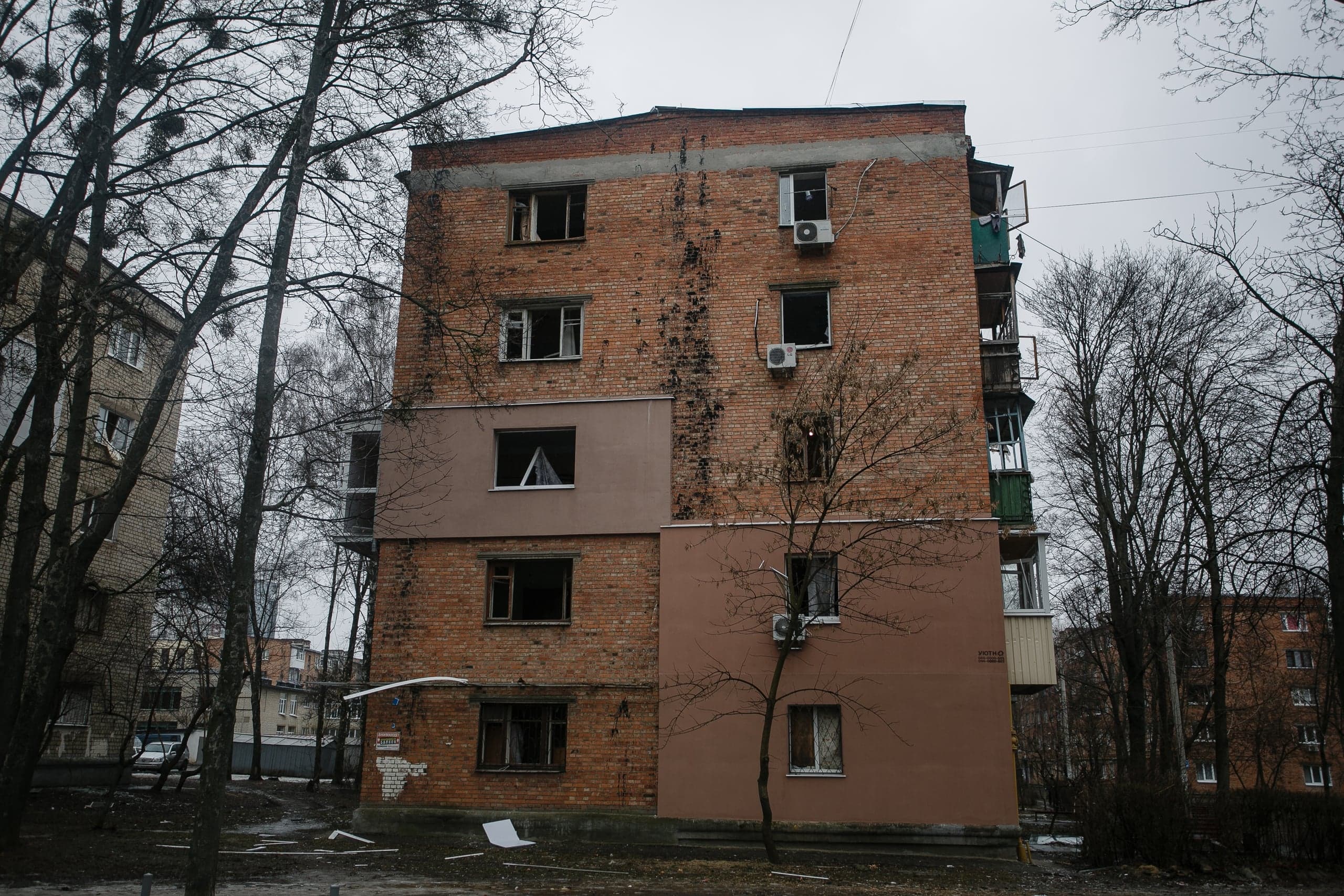 Поврежденный кирпичный жилой дом с пустыми окнами и вертикальными следами гари на фасаде, в окружении голых деревьев. Пасмурная погода.